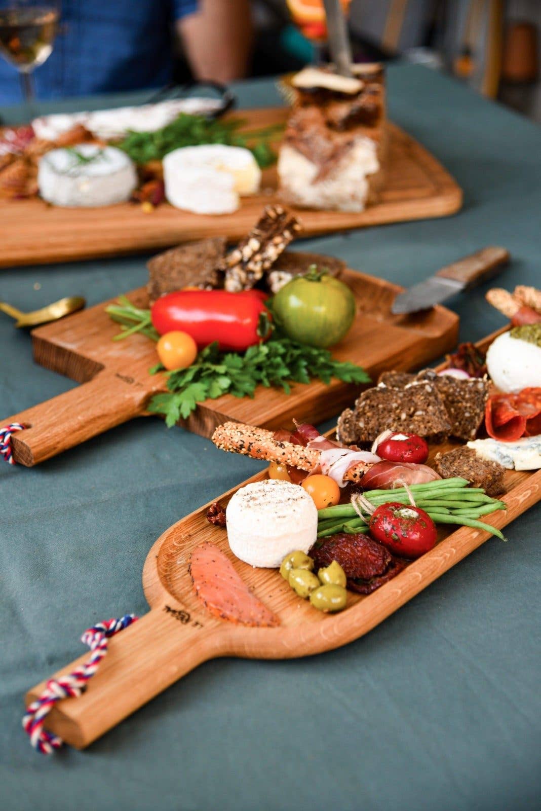 A LE RÉGAL - Lange Schaufel – großes Aperitifbrett aus Eichenholz mit Käse, Crackern, Brot, Kräutern, Oliven und Gemüse steht auf einem grünen Tischtuch. Im Hintergrund sind ein weiteres Brett, eine Person und eine nachhaltige Produktion zu erkennen.
