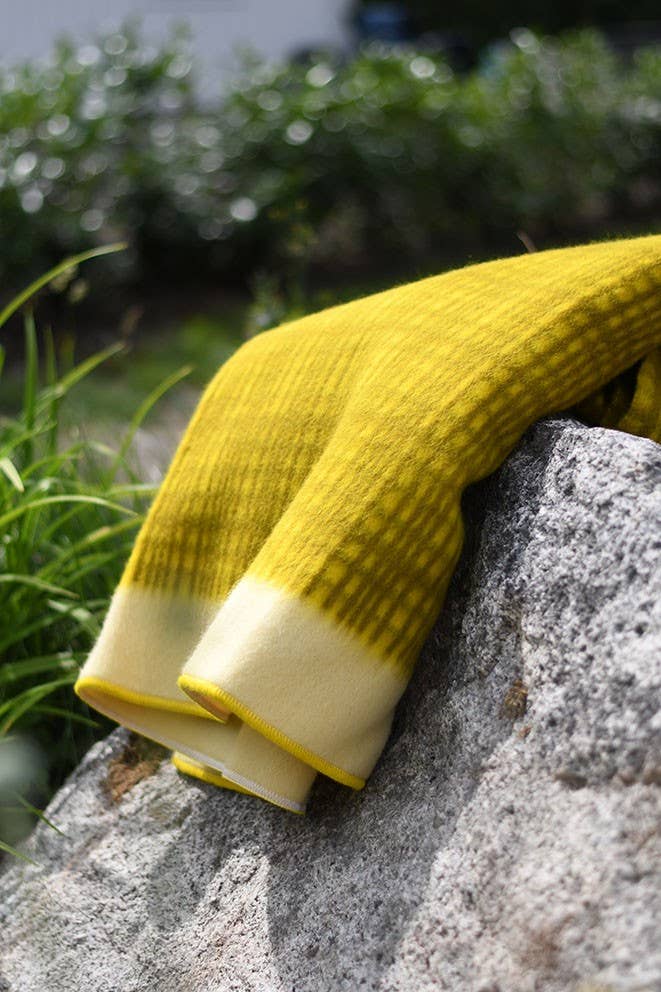 Die Darling Tagesdecke aus Lammwolle in Gelb von Sofie Sjöström Design liegt über einem großen grauen Stein im Freien, umgeben von grünen Pflanzen und unscharfem Blattwerk im Hintergrund.