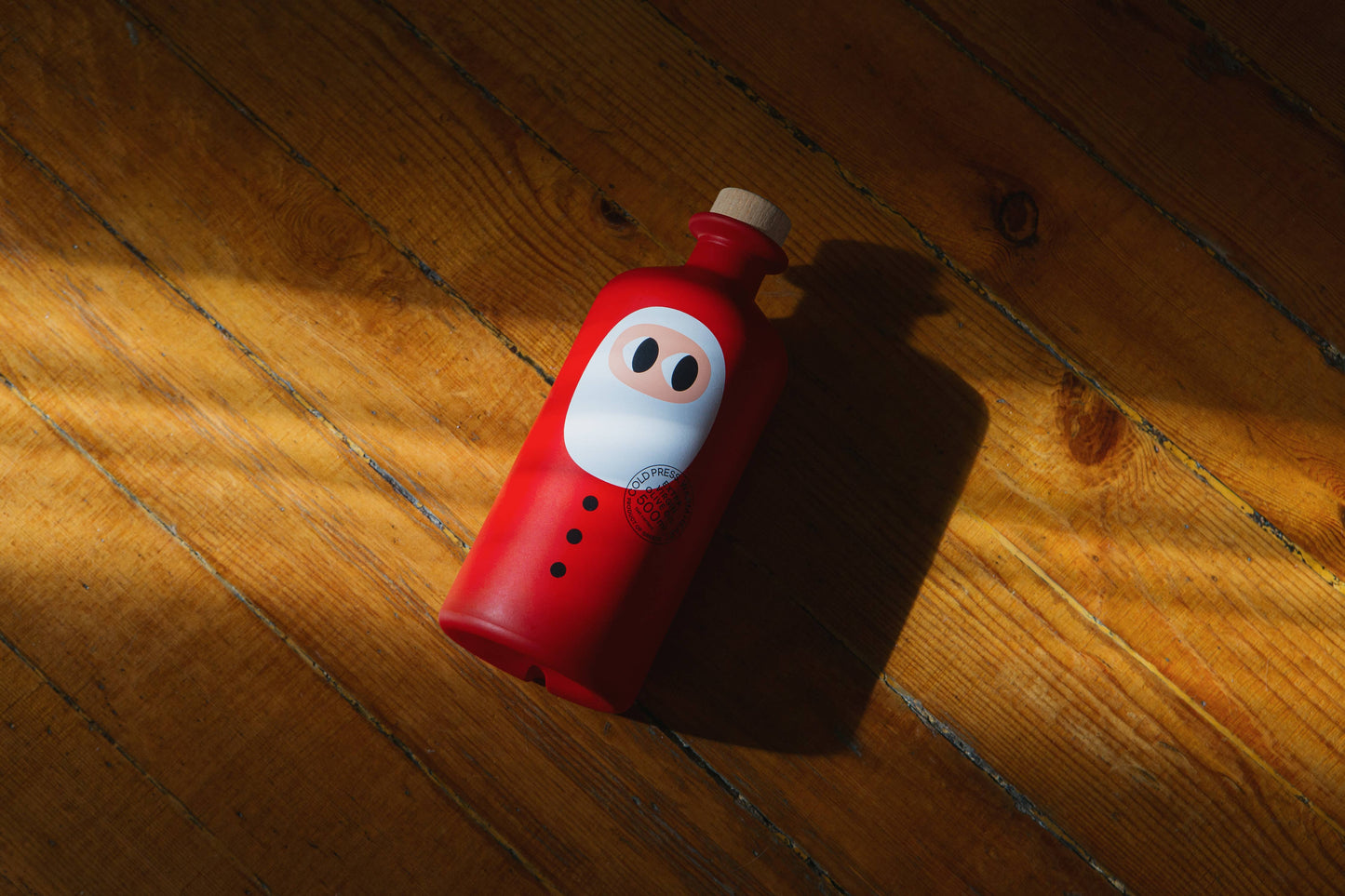 Eine 500 ml yiayia and friends - Natives Olivenöl extra Weihnachtsedition mit Holzkappe und Cartoon-Ninja-Gesicht ruht auf einem sonnenbeschienenen Boden - perfekt als Geschenk für Feinschmecker.