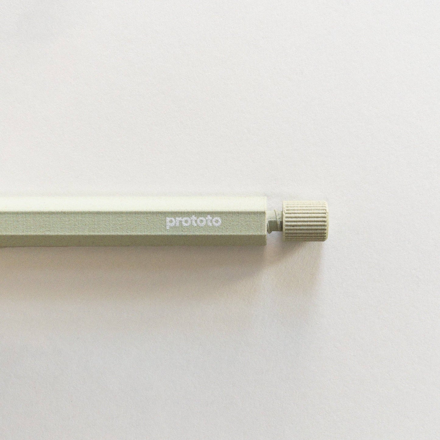 Eine Nahaufnahme des Prototo – Zero Waste Kugelschreibers in Salbei von Prototo, mit einem klimaneutralen Metallgehäuse, „Prototo“-Branding an der Seite und einem strukturierten runden Druckknopf auf einer weißen Oberfläche.