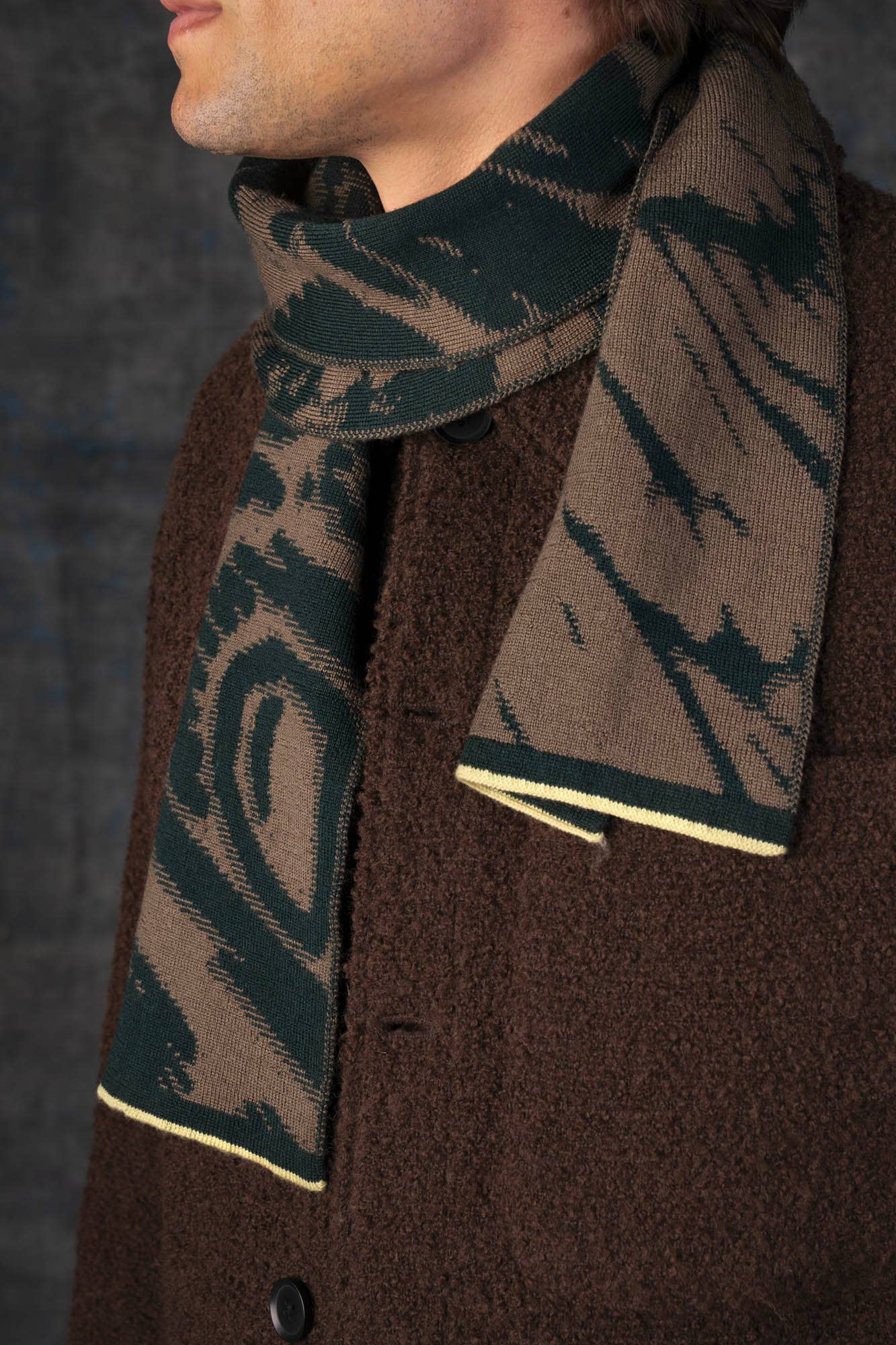 Eine Person in einem strukturierten braunen Mantel und dem "Schal aus Merinowolle - Furcula Wald Mini" von Wolvis mit dunkelgrünen und braunen Mustern sowie gelber Umrandung steht vor einem dunklen Hintergrund. Ihr Gesicht ist teilweise außerhalb des Rahmens.