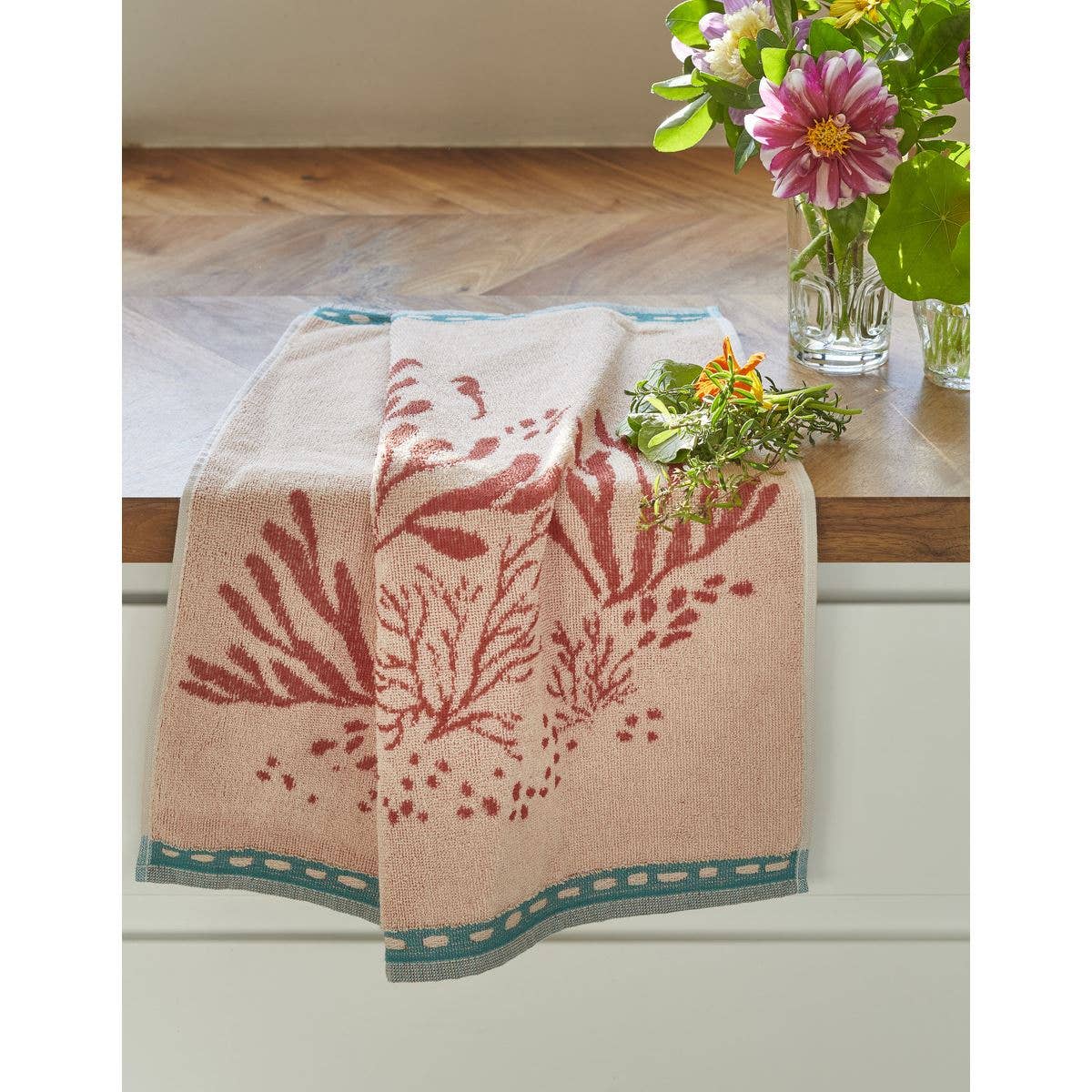 Ein Sylvie Thiriez | Handtuch Océan 50x50 mit rotem Korallenmuster und blaugrünem Rand ist über einer weißen Schublade drapiert. Daneben stehen auf der hölzernen Arbeitsplatte zwei Vasen aus klarem Glas, die mit bunten, frischen Blumen gefüllt sind.