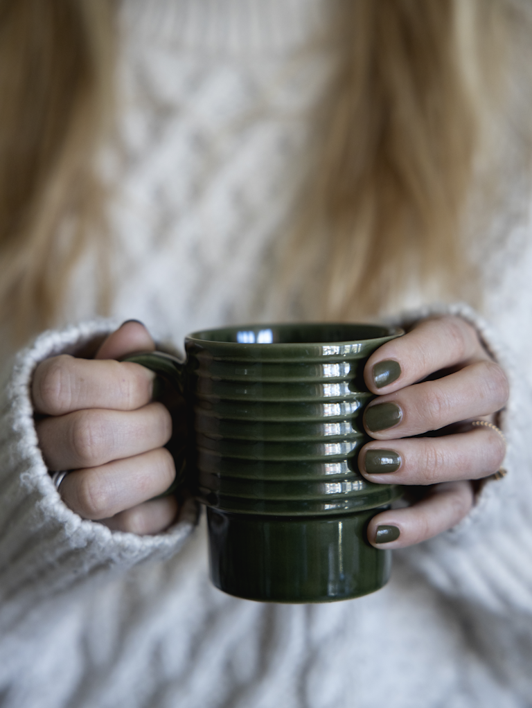Eine Person in einem weißen Strickpullover hält einen dunkelgrünen Sagaform GmbH Coffee & More Kaffeebecher Grün, 2-tlg mit passenden grünen Nägeln und schafft eine gemütliche Atmosphäre.