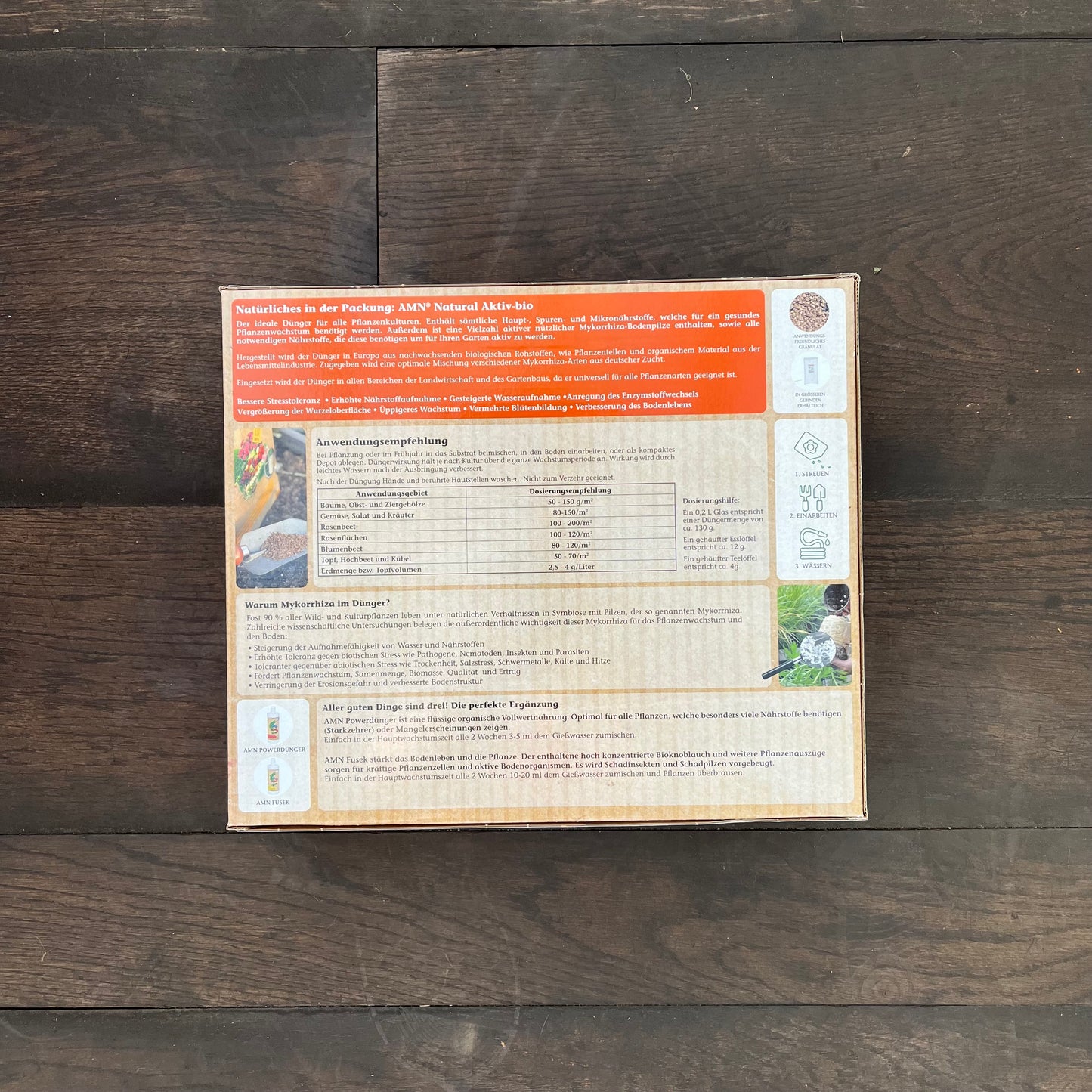 Auf der Rückseite einer AMN Natural Aktiv-bio-Verpackung finden sich deutscher Text, Nährwertangaben und Anwendungshinweise. Auf dunklem Holzboden zeigen kleine Bilder Pflanzen und Gartenarbeit und weisen die Packung als AMNs Gesunddünger mit Mykorrhiza aus.