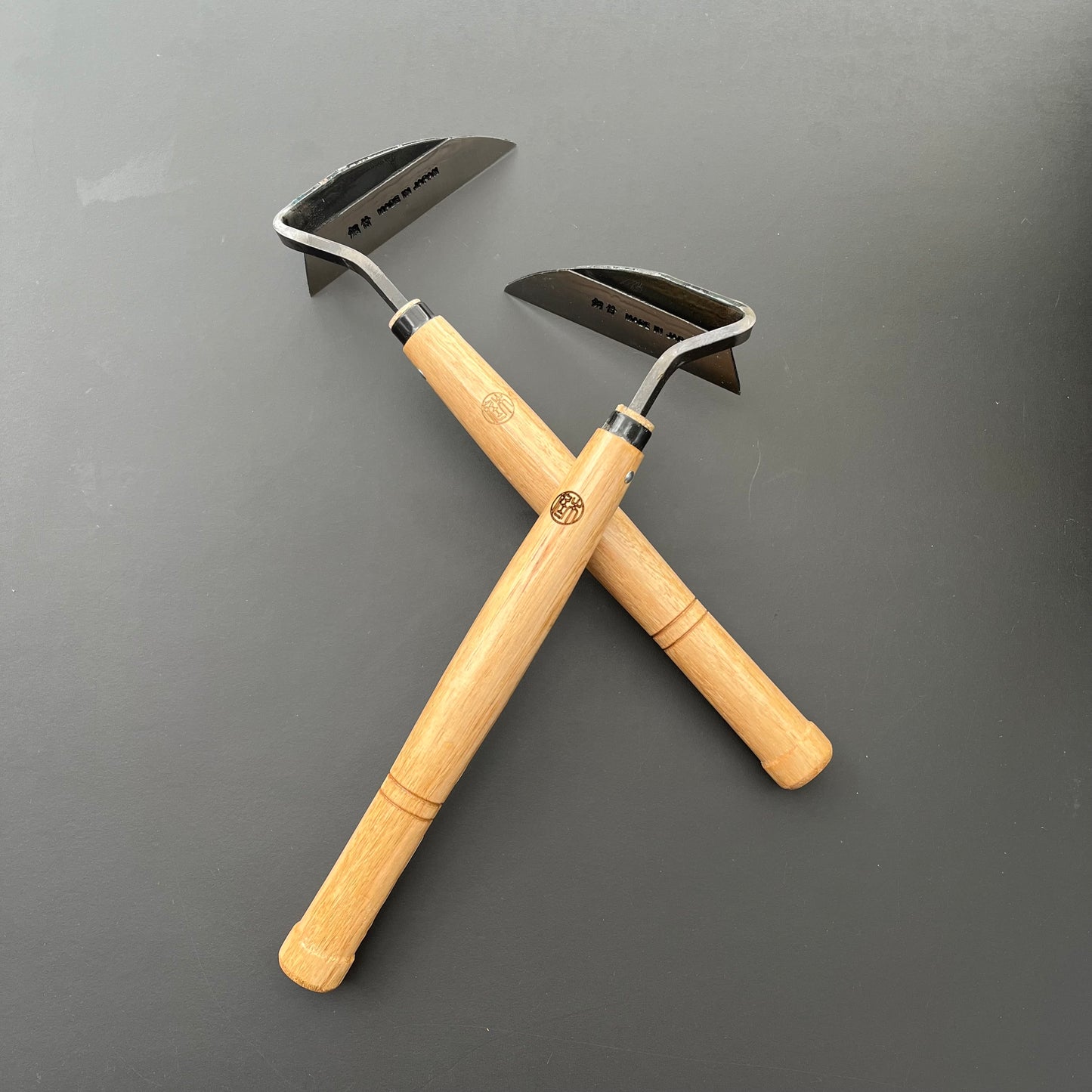 Zwei Niwaki-Unkrauthacken mit Holzgriffen und gebogenen Metallklingen sind in X-Form auf einer dunklen Oberfläche angeordnet – ergonomisch gestaltet von Niwaki für eine effektive Unkrautentfernung.