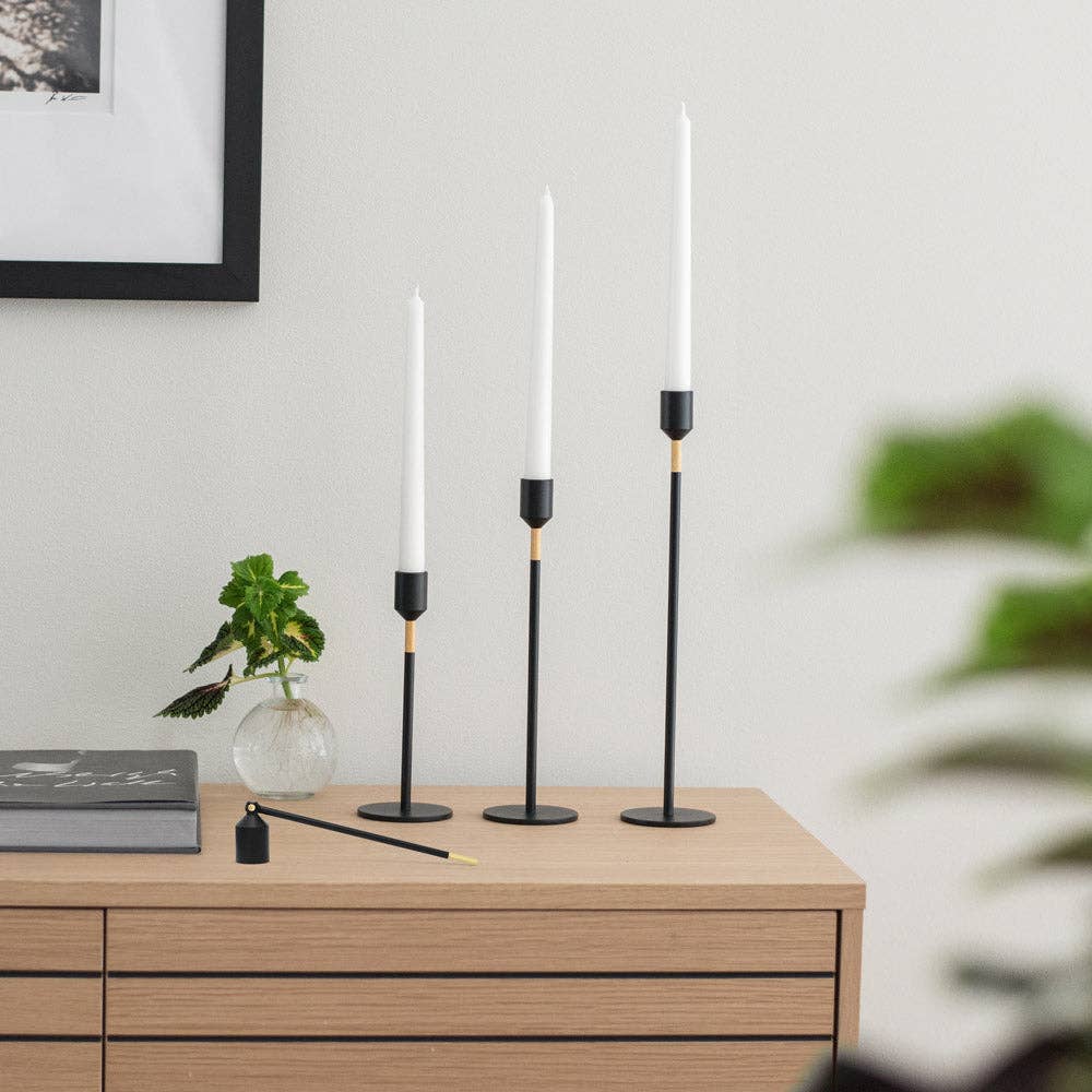 Auf einem hölzernen Sideboard sind drei Solstickan Design - Lichtlöscher - Schwarz Kerzenlöscher als Kerzenständer mit weißen Kerzen, eine kleine Glasvase mit einer grünen Pflanze, ein geschlossenes Buch und goldene Akzente auf dem gerahmten Bild darüber ausgestellt.