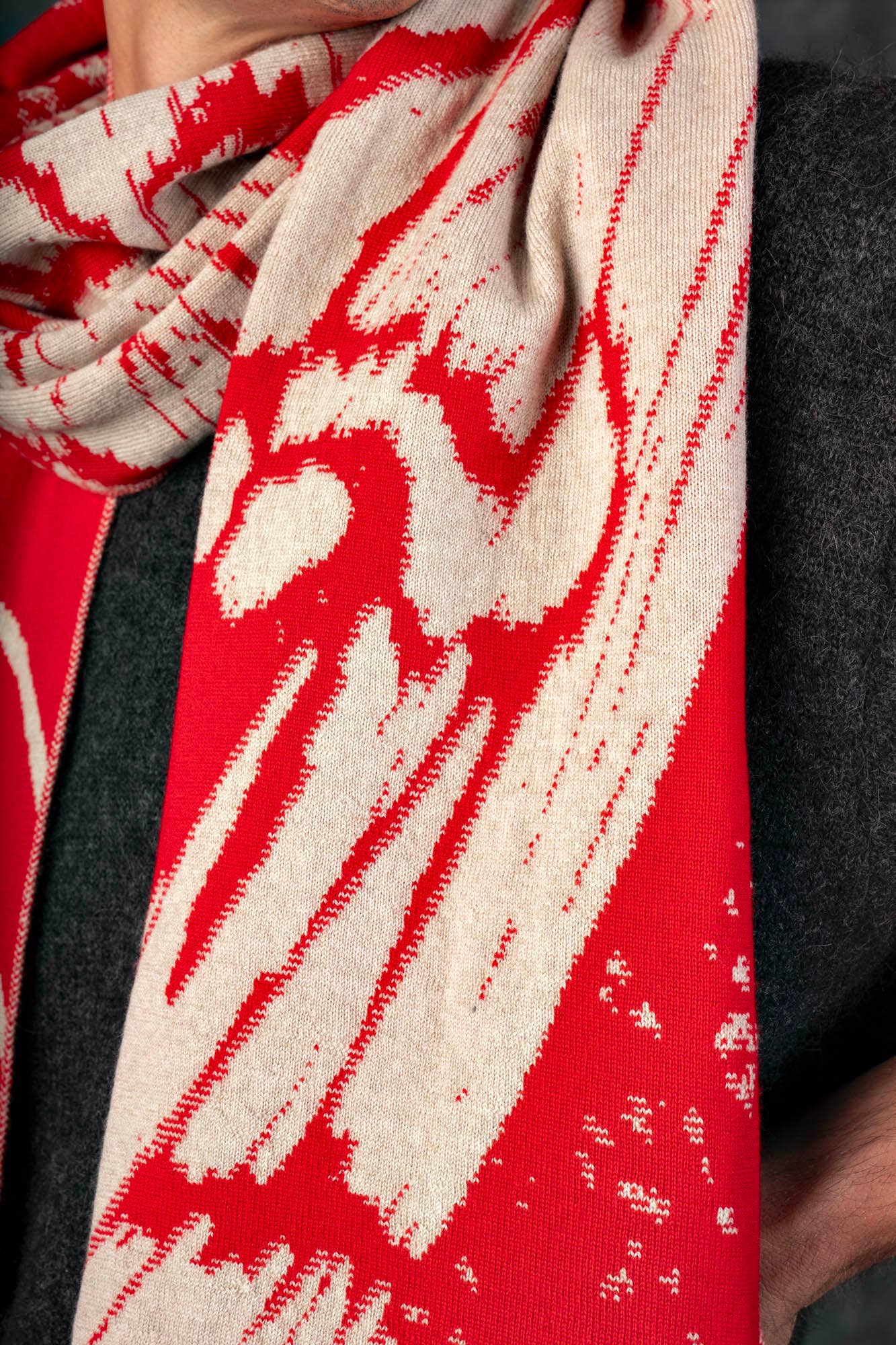Die Person trägt ein dunkelgraues Oberteil und drapiert den Wolvis Schal aus Merinowolle - Aurata Mohn Midi, der mit kühnen abstrakten Motiven in Rot und Beige, weicher Merinowolle und einem wendbaren Design aufwartet, das seine detaillierten Strickstrukturen hervorhebt.
