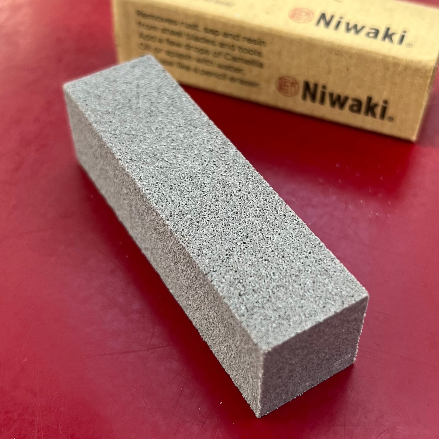 Ein rechteckiger, grauer Niwaki Mini Crean Mate mit grober Struktur ruht auf einer roten Fläche, mit einer braunen Niwaki-Box im Hintergrund, die auf seine Verwendung zum Schärfen von Werkzeugen hinweist.