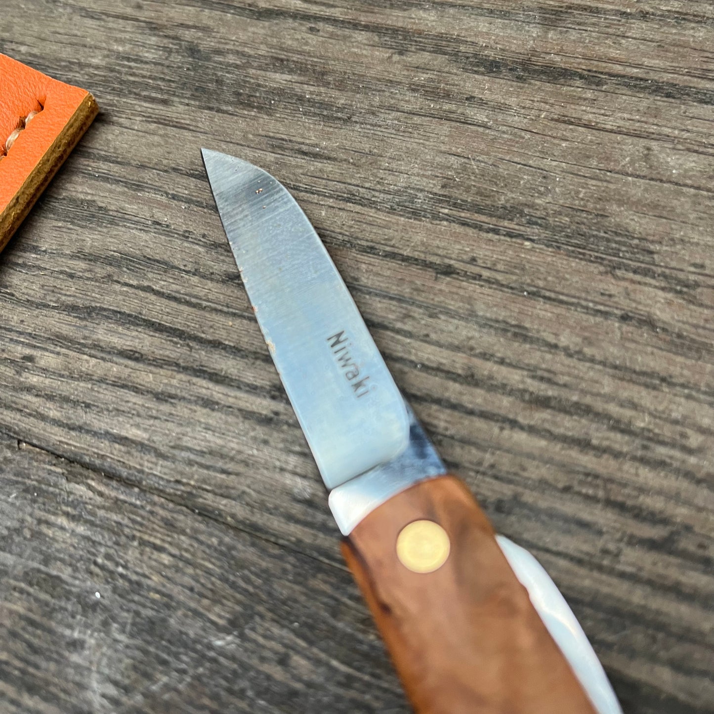 Ein Niwaki Kotoh X Niwaki Messer mit Holzgriff und teilweise geöffneter Klinge liegt auf einer verwitterten Holzoberfläche. Auf der Klinge ist „Train“ eingraviert, und in der oberen linken Ecke befindet sich eine Lederscheide.