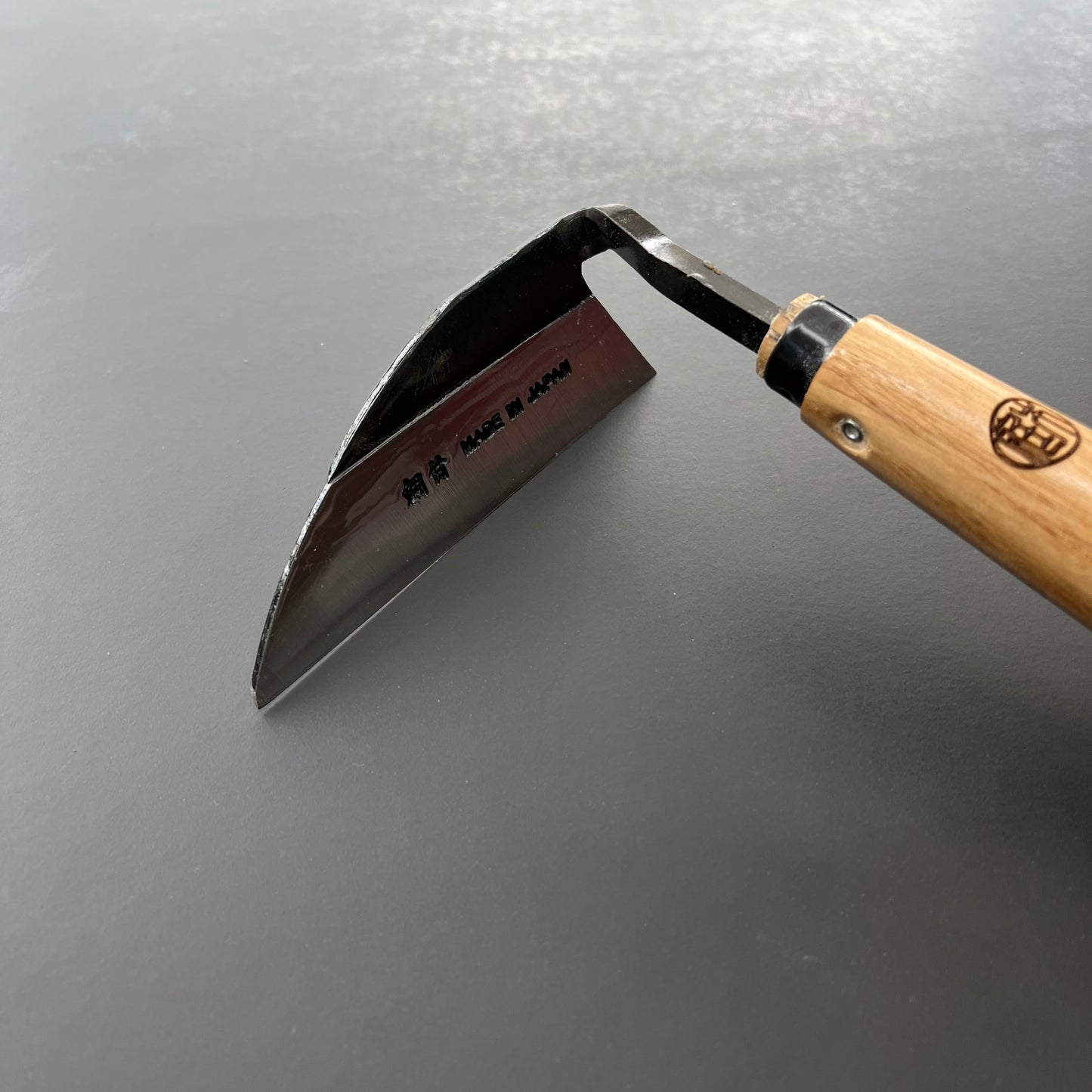 Eine Niwaki-Unkrauthacke, gefertigt von Niwaki zum Entfernen von Unkraut, liegt auf einer dunklen Oberfläche. Die gebogene Metallklinge mit eingravierten japanischen Schriftzeichen und dem Schriftzug „MADE IN JAPAN“ ist mit einem Holzgriff für ergonomisches Design kombiniert.