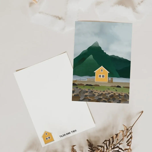Zwei Tilda&Theo - Postkarte - Norwegen Reise liegen auf einem hellen Untergrund: Eine zeigt ein gelbes Haus am Seeufer mit grünen Bergen, die andere ein kleines Hausmotiv und unten „Tilda&Theo“. Beide sind auf hochwertigem Naturpapier; daneben liegen getrocknete Blätter.