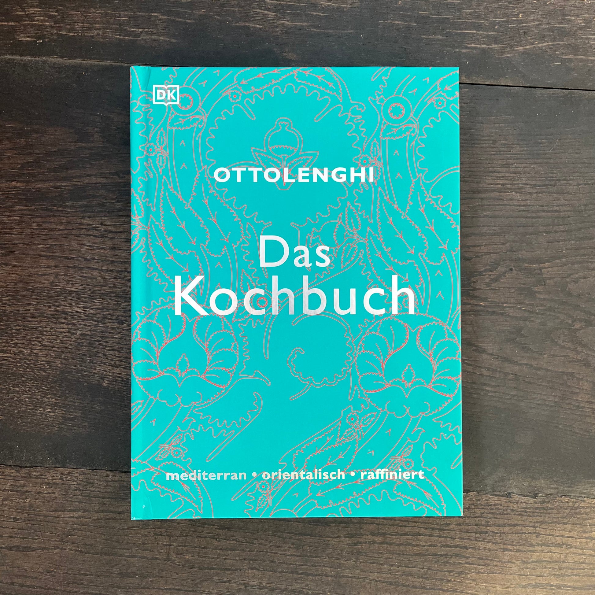 Auf einer dunklen Holzoberfläche liegt ein blaugrünes gebundenes DK Kochbuch - Ottolenghi „Das Kochbuch“, dessen Einband mit einem dezenten Muster und den Worten mediterran • orientalisch • raffiniert am unteren Rand verziert ist.
