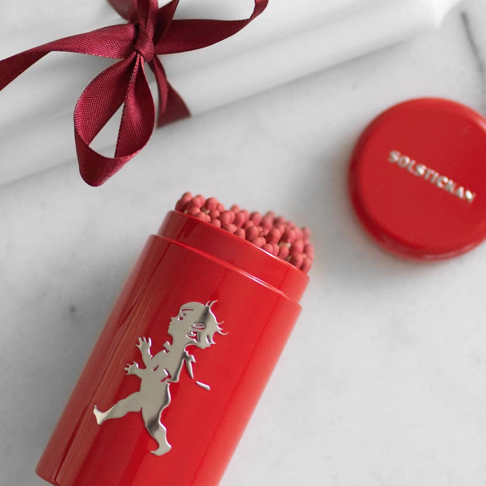 Ein roter Solstickan Design - Passender Schlauch - Rot (nur EU) von Solstickan Design mit einer silbernen Solsticke Boy-Silhouette hält extralange Streichhölzer mit rosa Spitzen. Der rote Deckel ist abgenommen, dahinter sitzt eine weiße Kerze mit burgunderrotem Band.