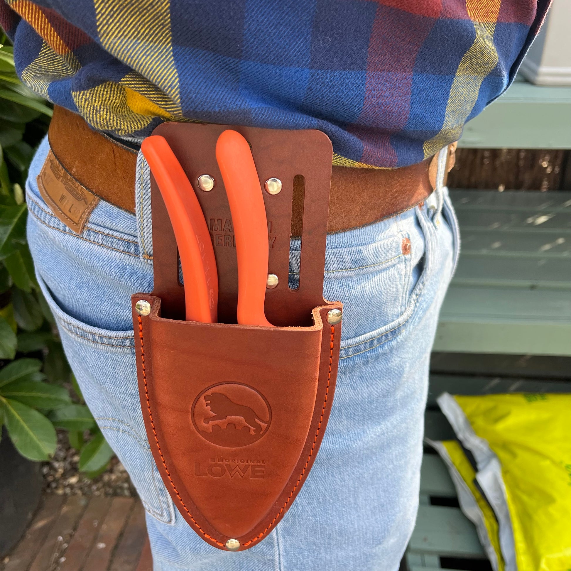 In blauer Jeans, kariertem Hemd und braunem Gürtel trägt eine Person zwei orangefarbene Werkzeuge in einem Original Löwe - Lederholster für Gartenscheren von Löwe, bereit für effiziente Gartenarbeit.