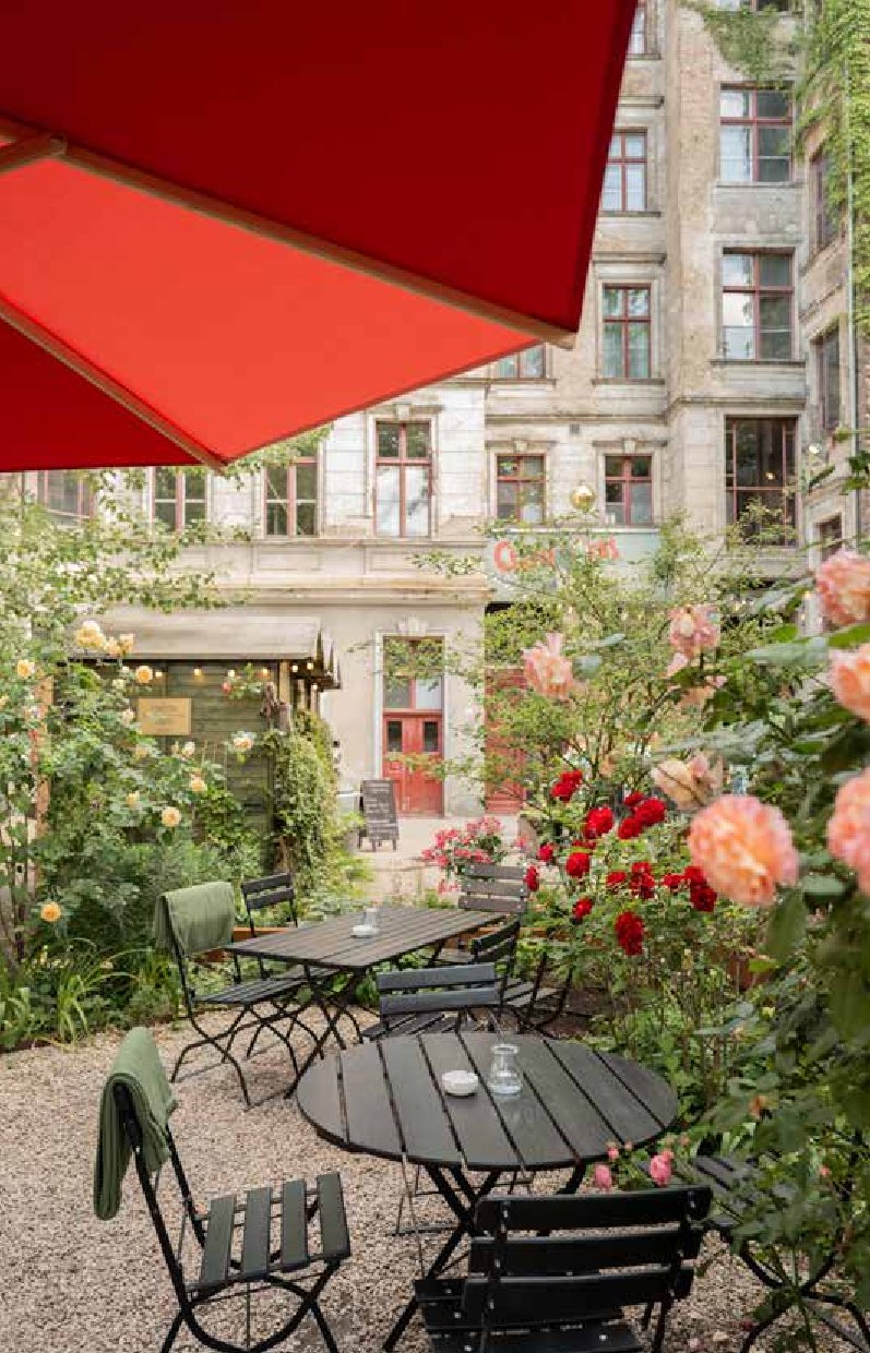 Genießen Sie eine gemütliche Gartencafé-Terrasse, wie sie in "Stadtoasen in Berlin" vom Bebra Verlag abgebildet ist, mit schwarzen Tischen und Stühlen inmitten von blühenden Blumen unter einem roten Sonnenschirm, vor einem alten Gebäude mit rot gerahmten Fenstern.
