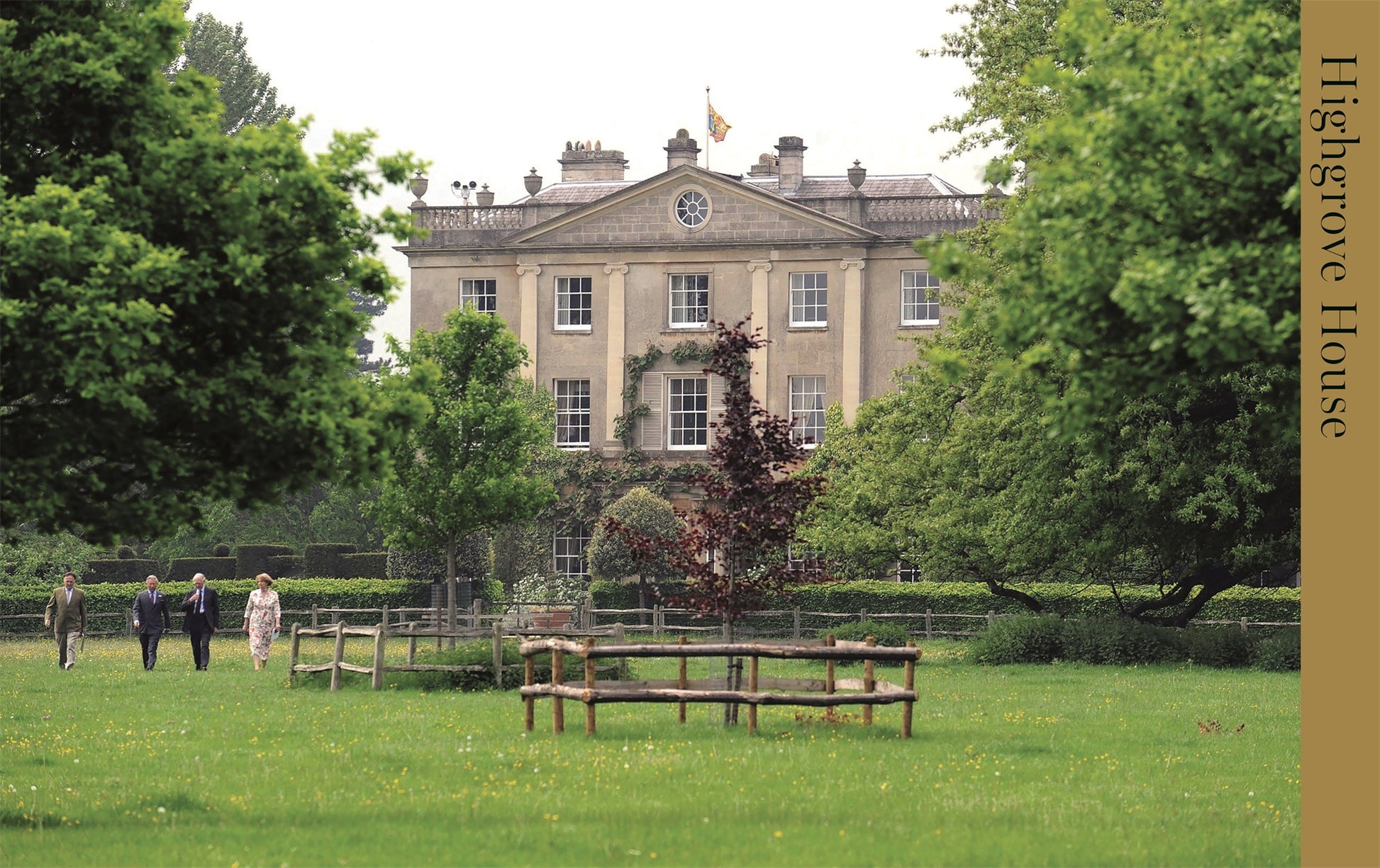 Vier Personen überqueren einen grasbewachsenen Rasen in Richtung des prächtigen Highgrove House, das für die Royal TeaTime des DK Verlags bekannt ist, die inmitten von Bäumen und Gärten die königliche Küche und britisches Gebäck zelebriert. Auf dem Rasen davor ist eine Holzkonstruktion zu sehen.