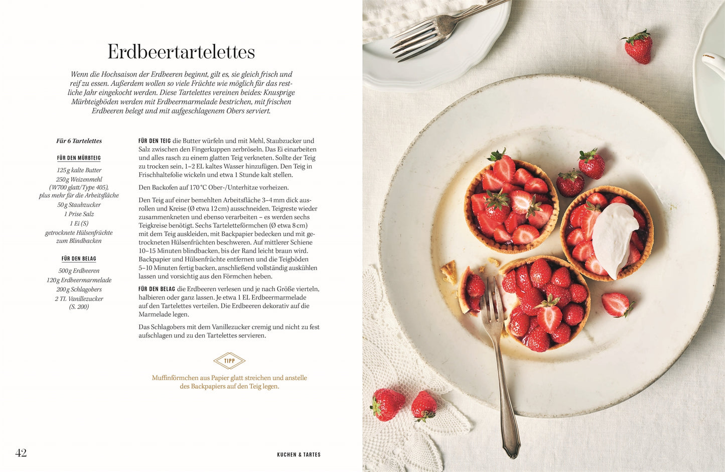 Ein weißer Teller mit drei Erdbeertörtchen aus der "Wiener Zuckerbäckerei" des DK Verlags steht auf einem hellen Tischtuch, umgeben von ganzen Erdbeeren und Besteck; links ist ein von der traditionellen österreichischen Küche inspiriertes Rezept zu sehen.
