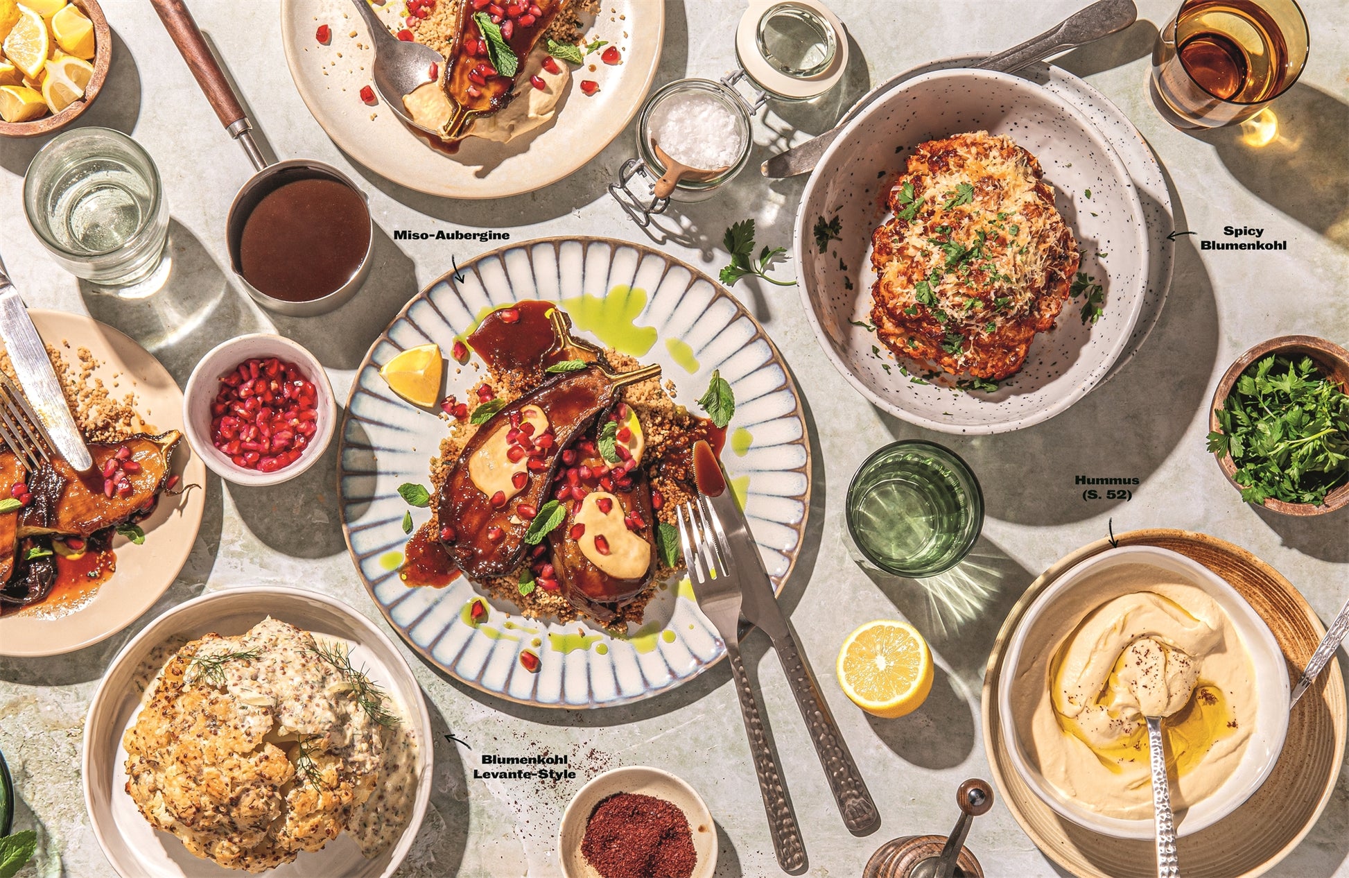 Ein gedeckter Tisch mit mediterranen Gerichten wie Hummus, würzigem Blumenkohl, Miso-Aubergine, Granatapfelkernen und frischen Kräutern aus „Mach’s lecker!“ vom DK Verlag, alles hübsch angerichtet auf weißen Tellern unter natürlichem Licht.