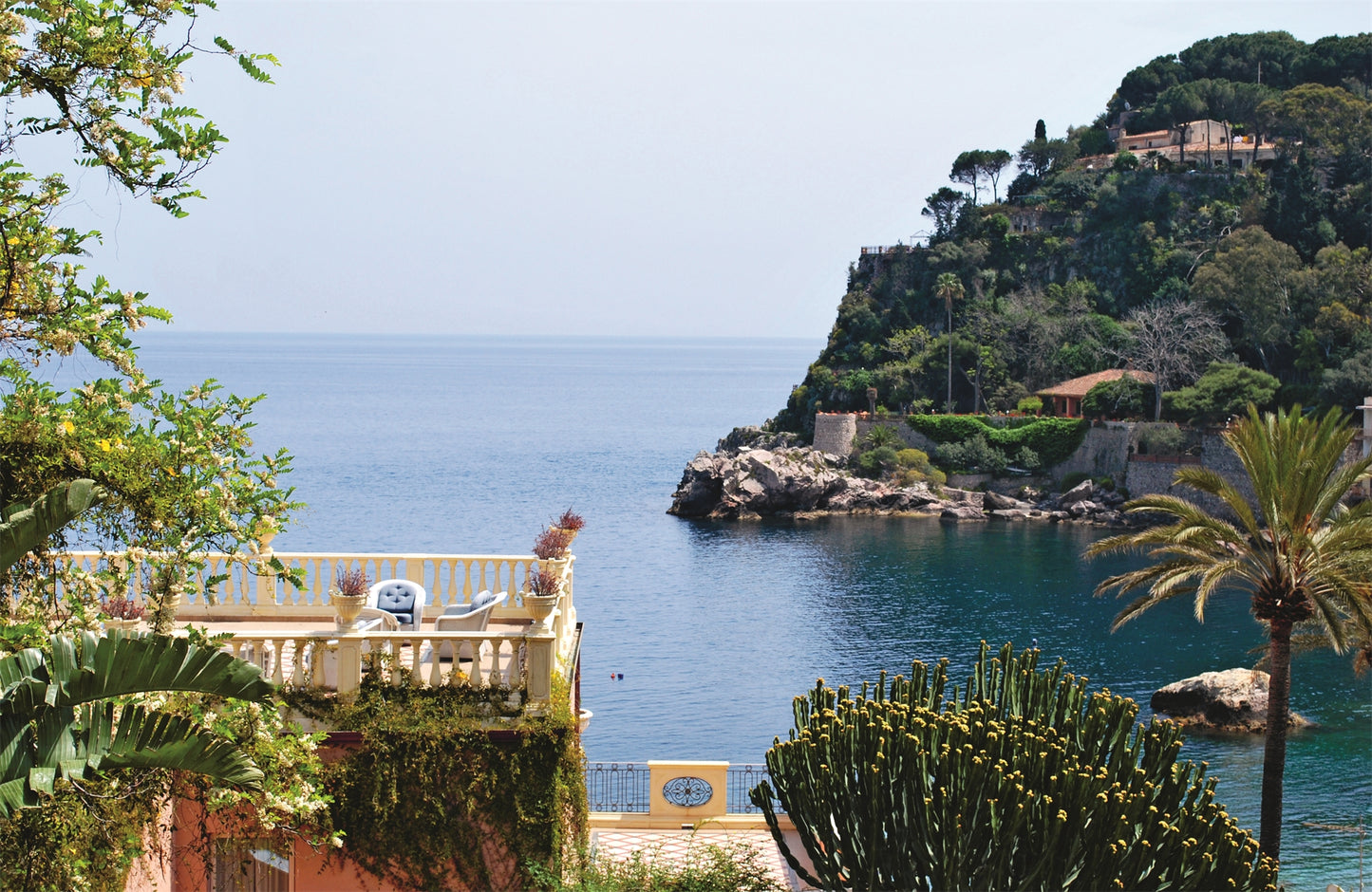 Erleben Sie die authentische sizilianische Atmosphäre von "Sizilien in meiner Küche" aus dem DK Verlag: eine gelbe Villa mit Terrasse mit Blick auf blaues Meer und üppige Hügel, umgeben von tropischen Pflanzen und Palmen.