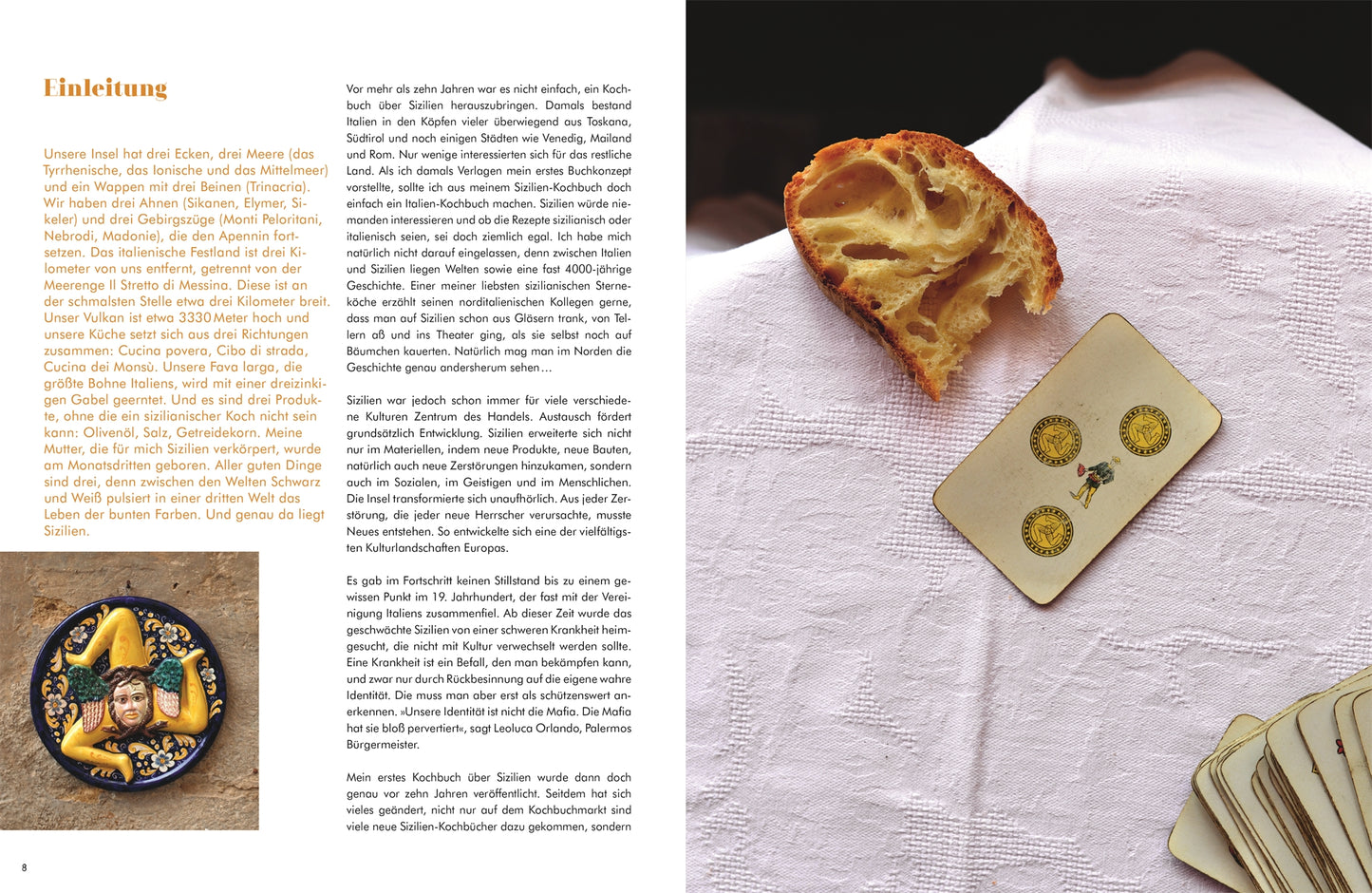 Eine dicke Scheibe Brot und eine Spielkarte liegen auf einem weißen Tuch. Daneben steht ein Teller mit gelben Mustern, auf dem Oliven und eine kleine Figur liegen - eine Szene, die von "Sizilien in meiner Küche" von Cettina Vicenzino aus dem DK Verlag inspiriert ist. Der Text erscheint auf der linken Seite.