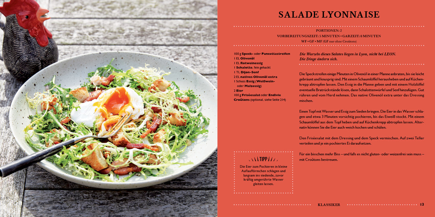 Eine Schüssel Salade Lyonnaise mit pochiertem Ei, Speck, Croutons und Käse steht neben dem Kochbuch „LEON. Hundert Salate.“ aus dem Dumont Verlag; im Hintergrund ist eine Keramikfigur eines Hahns zu sehen.