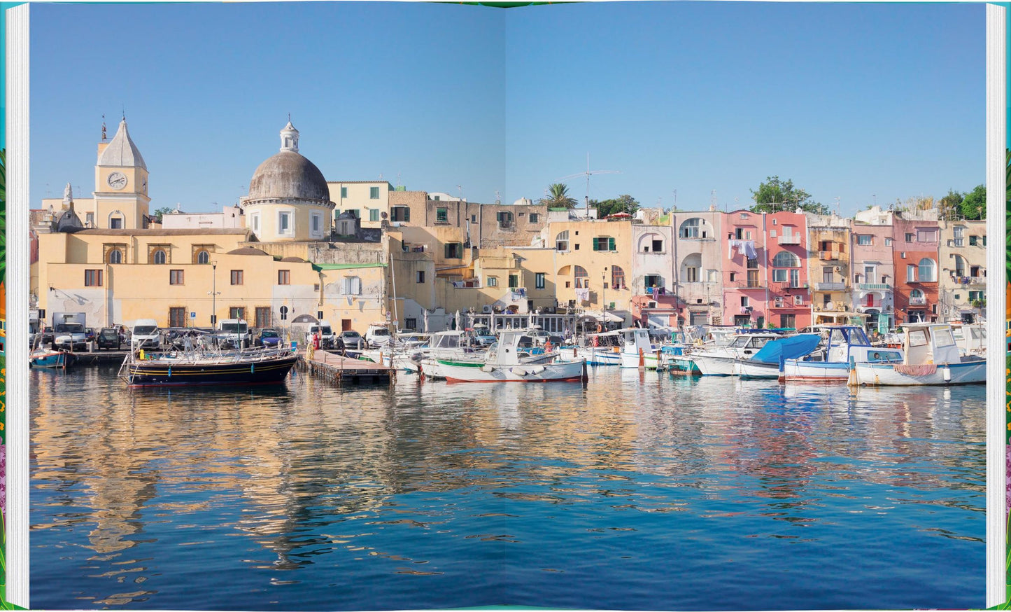 Bunte pastellfarbene Gebäude und eine Kirche säumen die Uferpromenade von Amalfi, wo Boote unter klarem Himmel auf blauem Wasser treiben – eine Szene, die perfekt zu „Die Küche von Amalfi“ vom Gerstenberg Verlag passt.