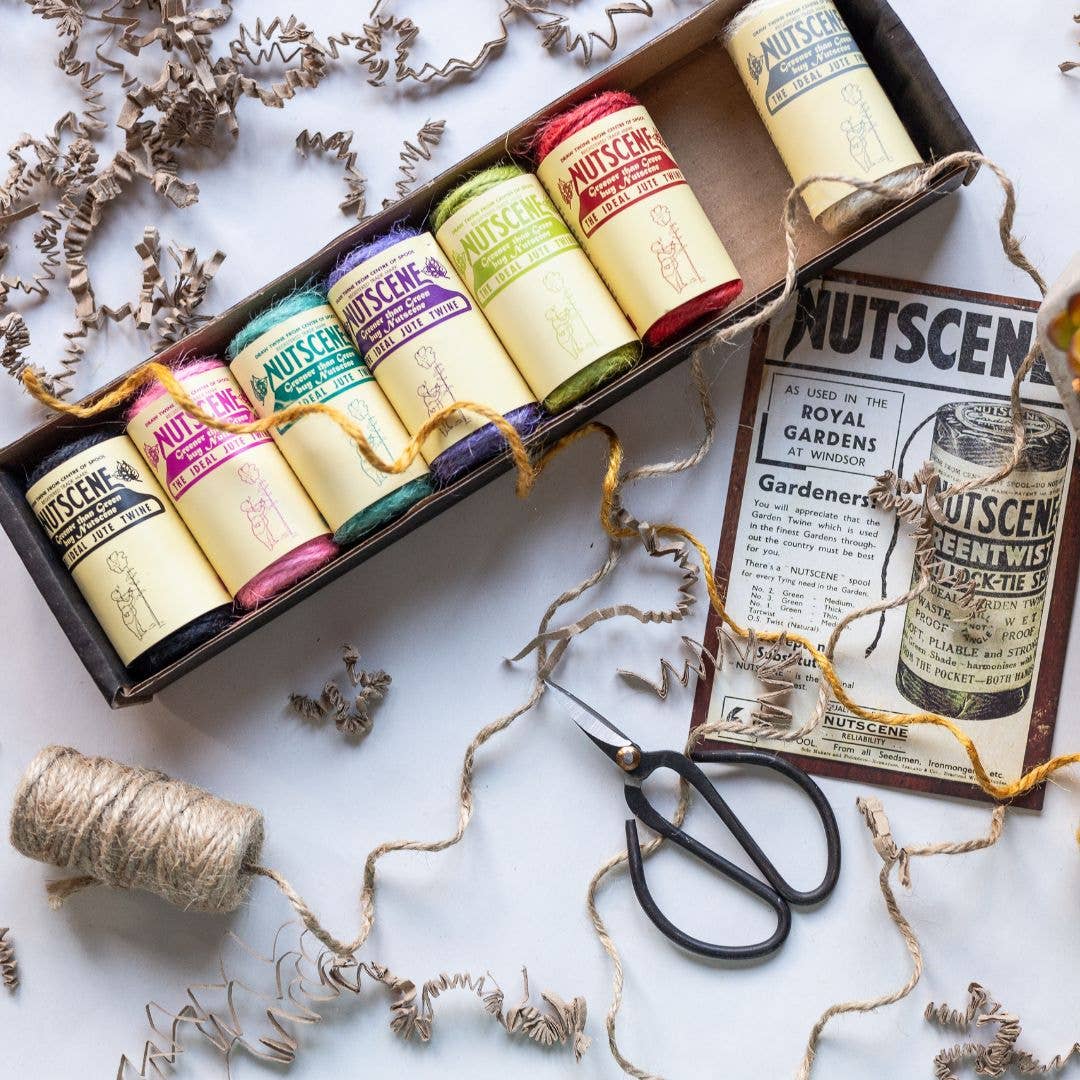 Ein bezauberndes Geschenkset von Nutscene Twines Ltd aus der Mini-Twines-Kollektion mit 8 Schnüren (je 42 Fuß) liegt auf weißem Untergrund, umgeben von einem Flyer, Schere, losem Garn und braunen Papierschnipseln.