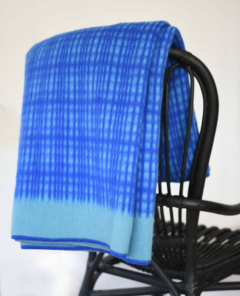 Die Darling Tagesdecke aus Lammwolle in Blau von Sofie Sjöström Design mit festem Rand ist über einen schwarzen Korbstuhl drapiert und steht vor einem schlichten weißen Hintergrund.