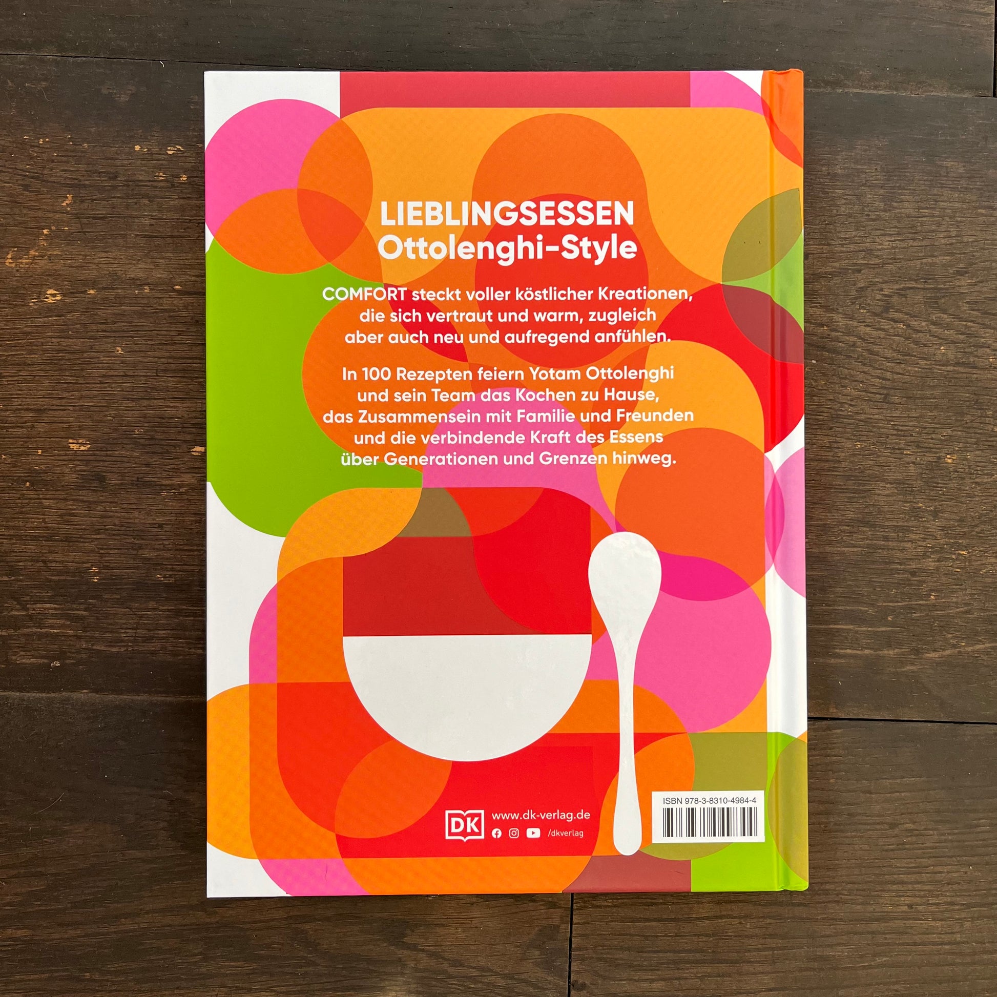 Die Rückseite des Kochbuchs „Comfort“ von DK – Ottolenghi zeigt leuchtende, sich überlappende Kreise in Orange, Rot, Pink und Grün mit fettem weißem deutschem Text und einer großen Löffelgrafik auf dunklem Holzhintergrund, wodurch die natürlichen Zutaten hervorgehoben werden.
