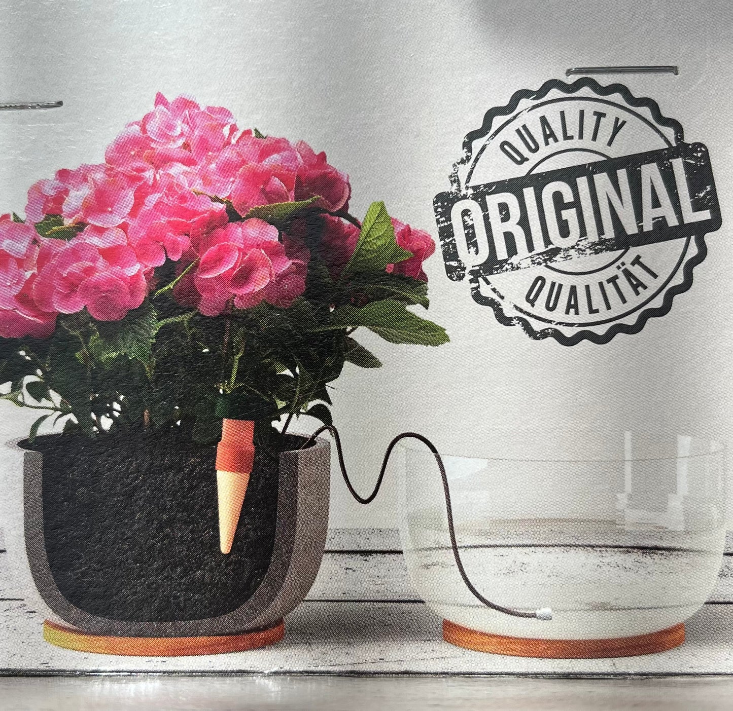 Eine rosa blühende Pflanze in einem schwarzen Topf ist durch einen Schlauch mit einer klaren Wasserschale verbunden und demonstriert das Blumat Classic für Zimmer- und Balkonpflanzen Bewässerungssystem von Blumat. Ein Siegel mit "ORIGINAL" und "QUALITY" steht neben der Einrichtung.