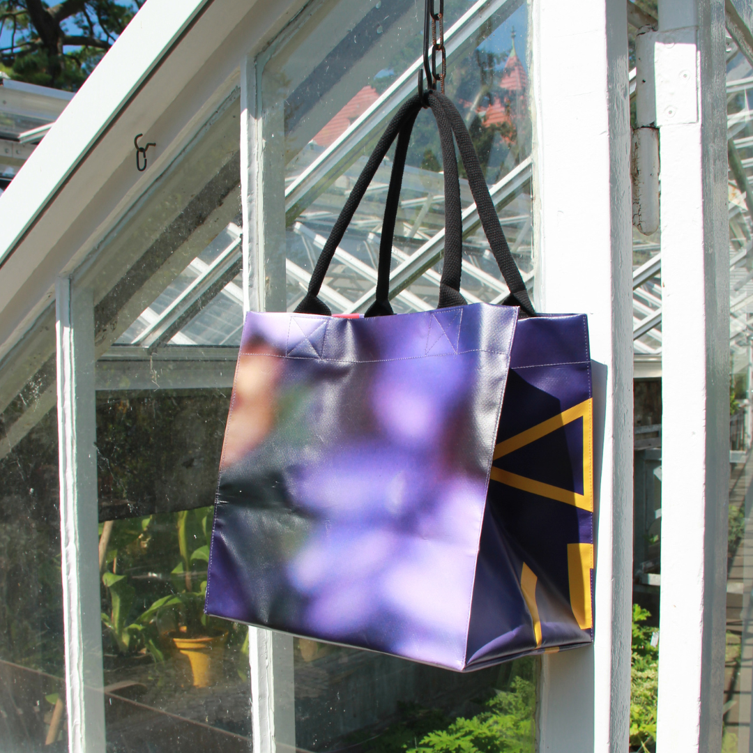 Die Upcycling KGA Tasche 'Aster' von der Königlichen Gartenakademie mit schwarzen Griffen hängt an einem Haken in der Nähe einer gläsernen Gewächshauswand, von der aus man das Sonnenlicht und die Grünpflanzen sehen kann.