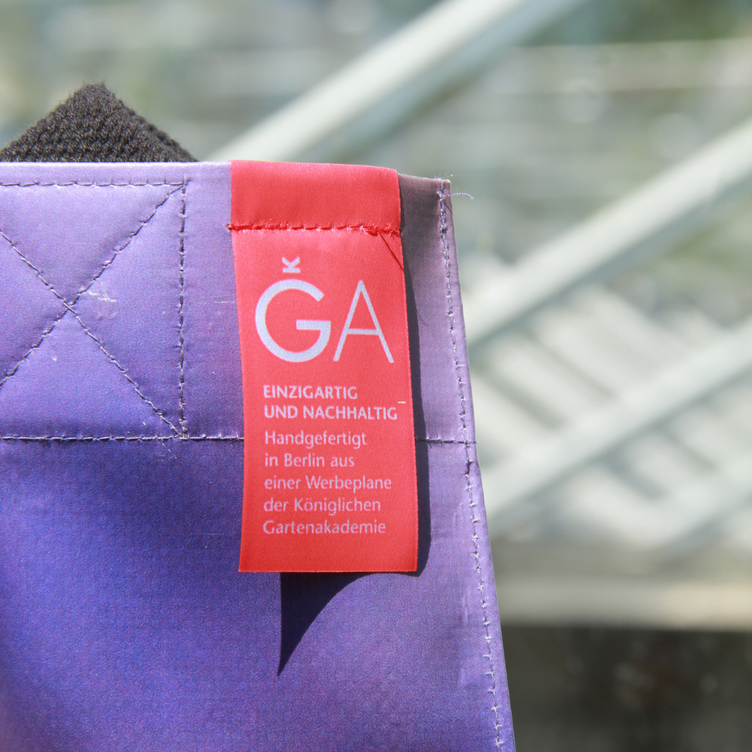 Eine Nahaufnahme der lilafarbenen Upcycling KGA Tasche 'Aster' von der Königlichen Gartenakademie, die ein rotes Schild mit weißem Text aufweist: "EINZIGARTIG UND NACHHALTIG Handgefertigt in Berlin aus einer Werbeplane.