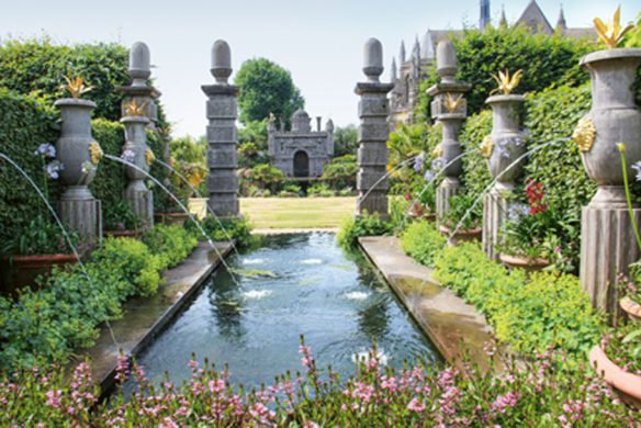 Die Ausstellung "Gärtnern wie Gott in England" des Callwey Verlags zeigt einen formalen Garten mit englischem Design, einem rechteckigen, spiegelnden Pool, goldverzierten Steinsäulen, Wasserfontänen, üppigem Grün und einem stattlichen Steingebäude zwischen Bäumen.