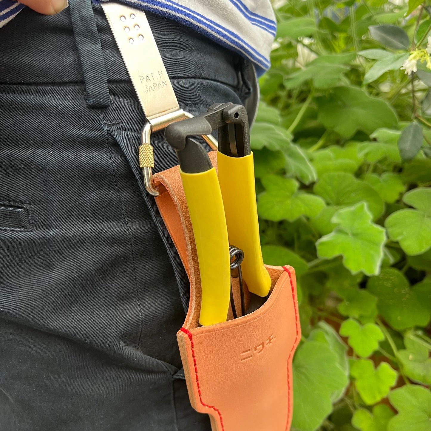 Eine Person in schwarzen Hosen hat einen Niwaki-Gürtelclip an ihrem Hosenbund befestigt und hält zwei Werkzeuge mit gelben Griffen, im Hintergrund ist grünes Laub zu sehen.