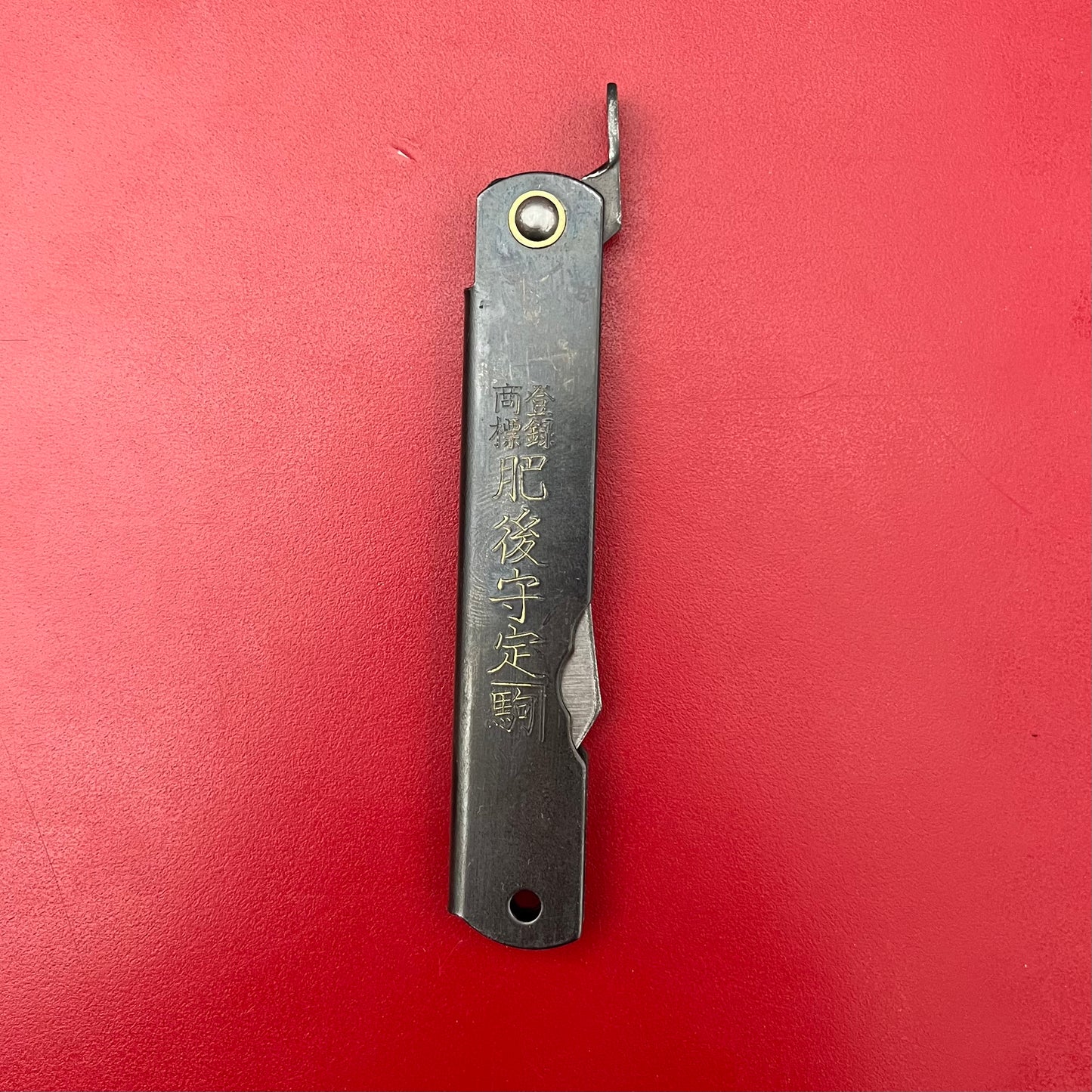 Das Niwaki SK Higonokami-Klappmesser von Niwaki ist geschlossen auf rotem Hintergrund abgebildet und verfügt über eingravierte Kanji, einen Metallgriff mit Niete und einen hervorstehenden Hebel für die scharfe Klinge.