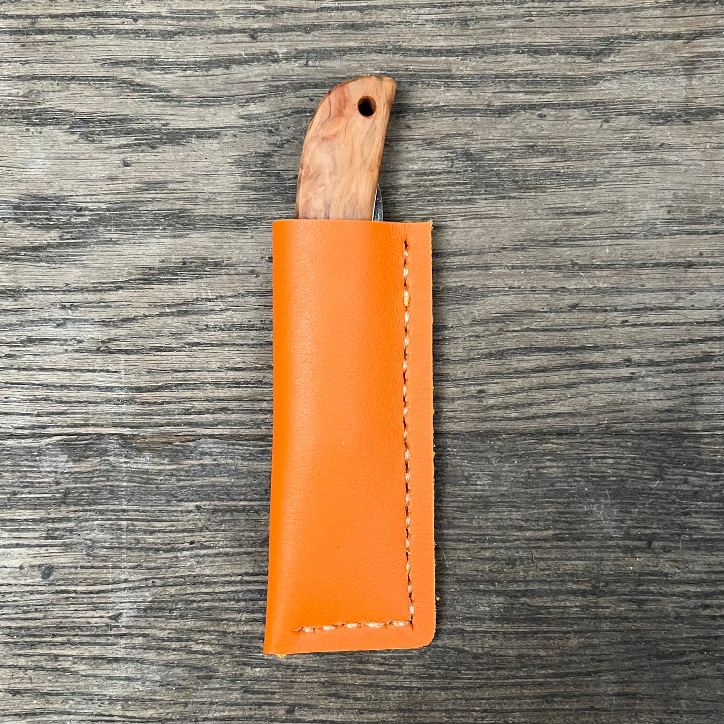 Ein Niwaki Kotoh X Niwaki-Messer mit Holzgriff liegt teilweise in einer orangefarbenen Lederhülle, wobei sich die leuchtende Scheide vom strukturierten Holz abhebt – von Niwaki für höchste Schneidleistung gefertigt.