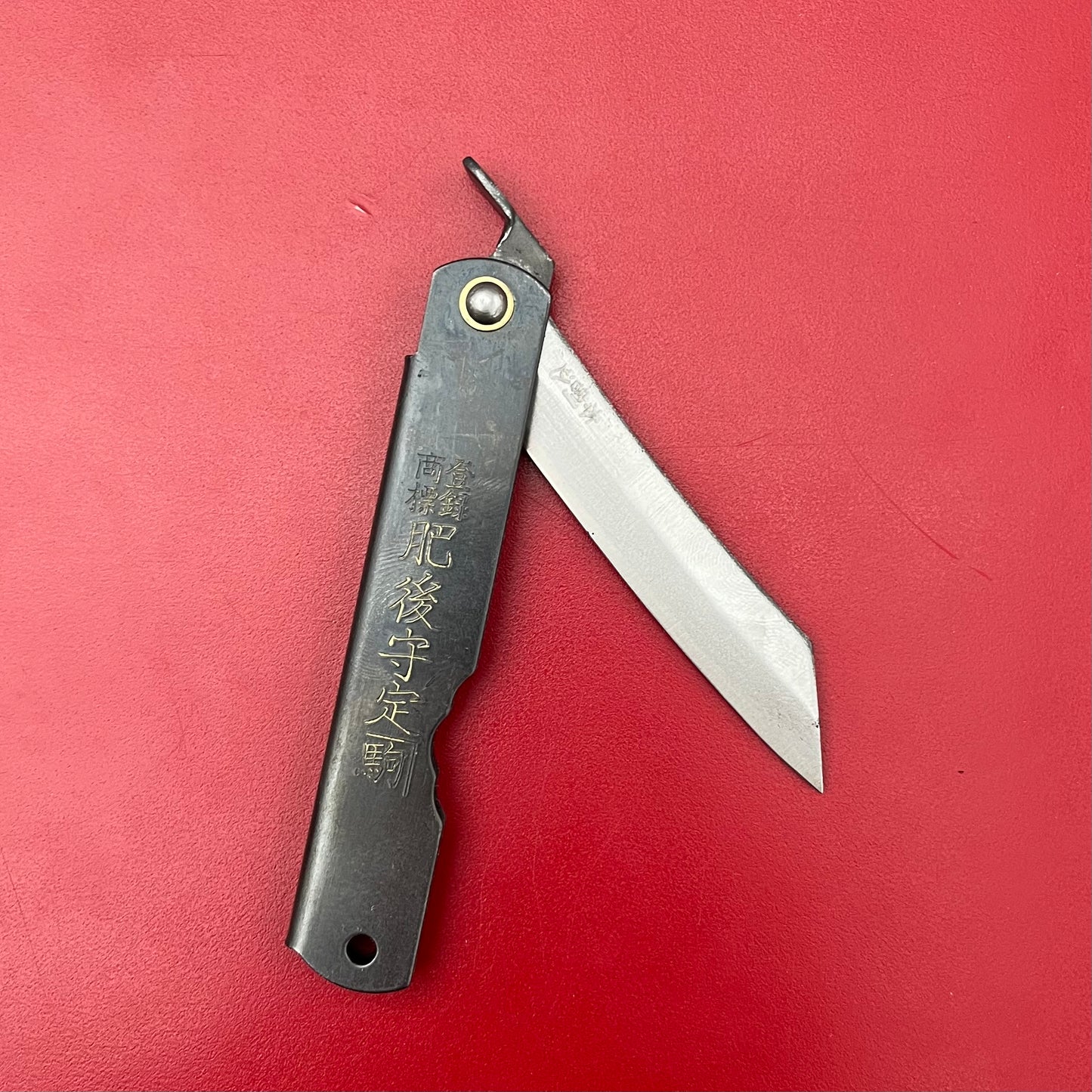 Auf einem einfarbig roten Hintergrund liegt ein Niwaki SK Higonokami-Klappmesser mit einer scharfen, teilweise geöffneten Klinge und einem schwarzen Griff mit eingravierten japanischen Schriftzeichen.