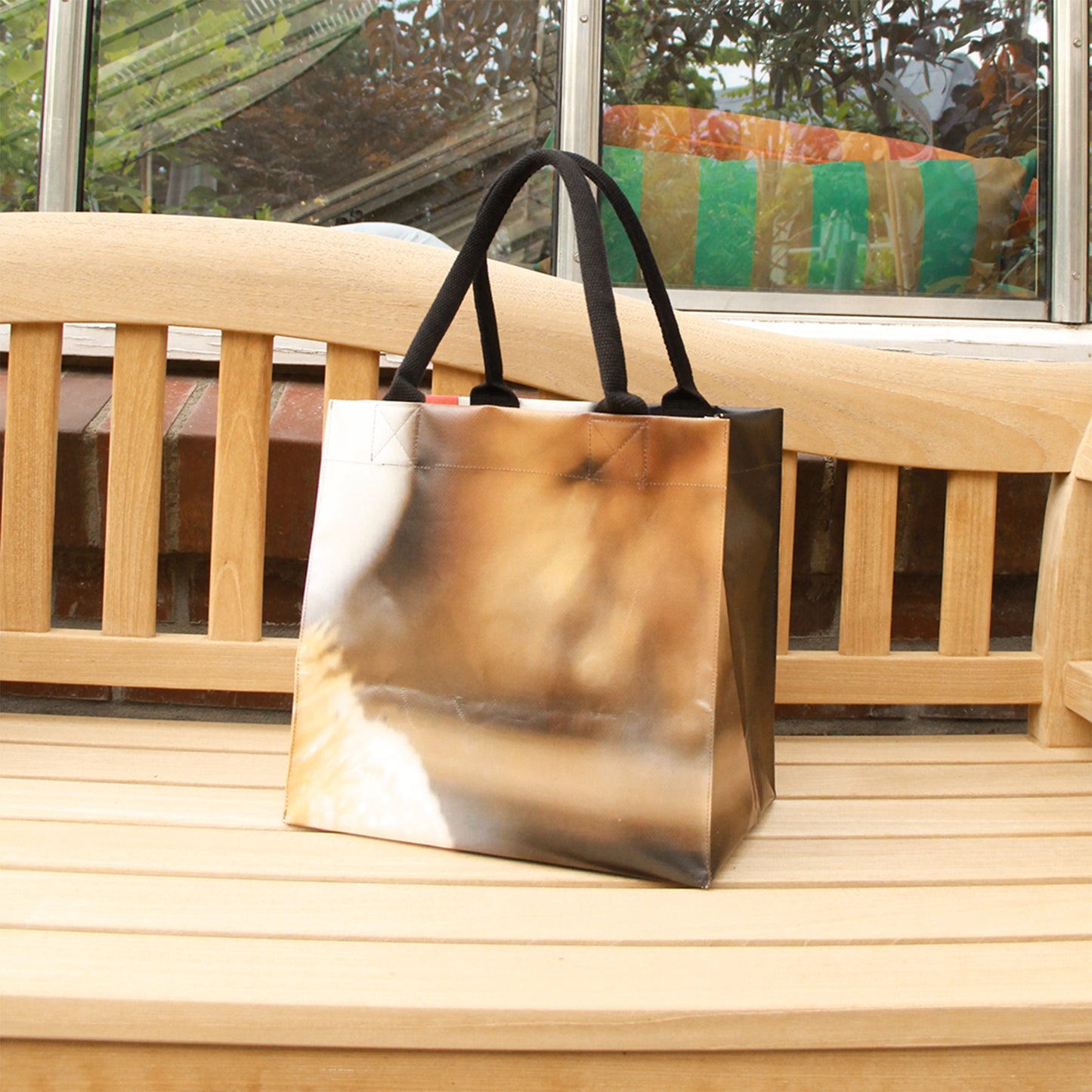 Die Upcycling KGA Tasche 'Torf' von Königliche Gartenakademie mit schwarzen Henkeln und abstraktem Print in Braun- und Beigetönen steht auf einer Holzbank im Freien, dahinter Fenster und bunte Kissen.