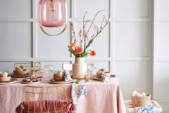 Das Esstischset Frühlingstraum des Callwey Verlags besteht aus einer rosafarbenen Tischdecke, goldenen Stühlen, rosafarbenen Tellern, Gläsern und einem floralen Tafelaufsatz - wunderschöne Dekorationsideen für das Osterfest. Eine rosafarbene Pendelleuchte vervollständigt den festlichen Look vor einer getäfelten Wand.