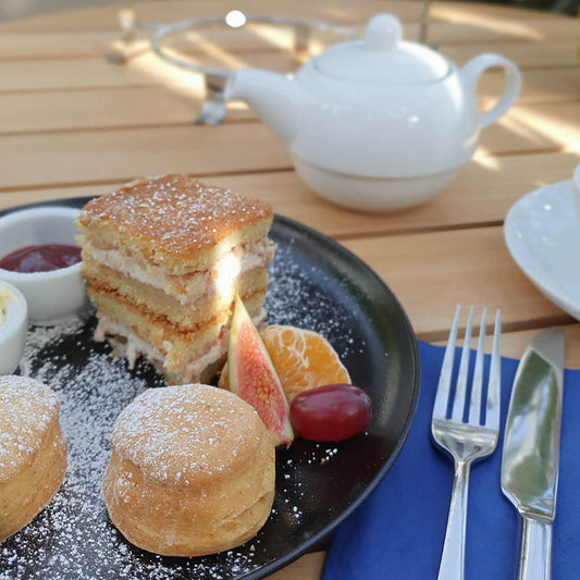 Genießen Sie den Di. 09.12.2025 | 14.30-16.00 AFTERNOON TEA - Christmas Special von Isabelle Van Groeningen: Scones, eine geschichtete Kuchenscheibe, Obst, Sahne und Marmelade auf einem schwarzen Teller, mit einer Teekanne und Teetasse - perfekt für eine festliche Teestunde.