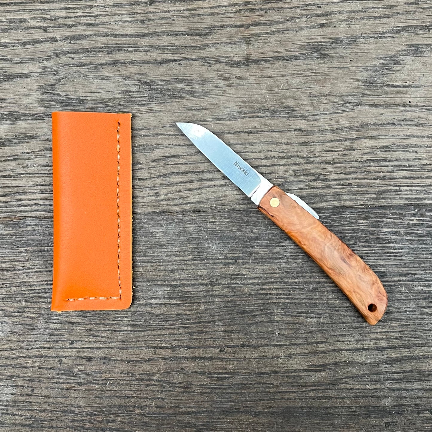 Ein kompaktes Kotoh X Niwaki-Messer von Niwaki mit Holzgriff liegt offen auf einer Holzoberfläche neben seiner orangefarbenen Lederscheide.