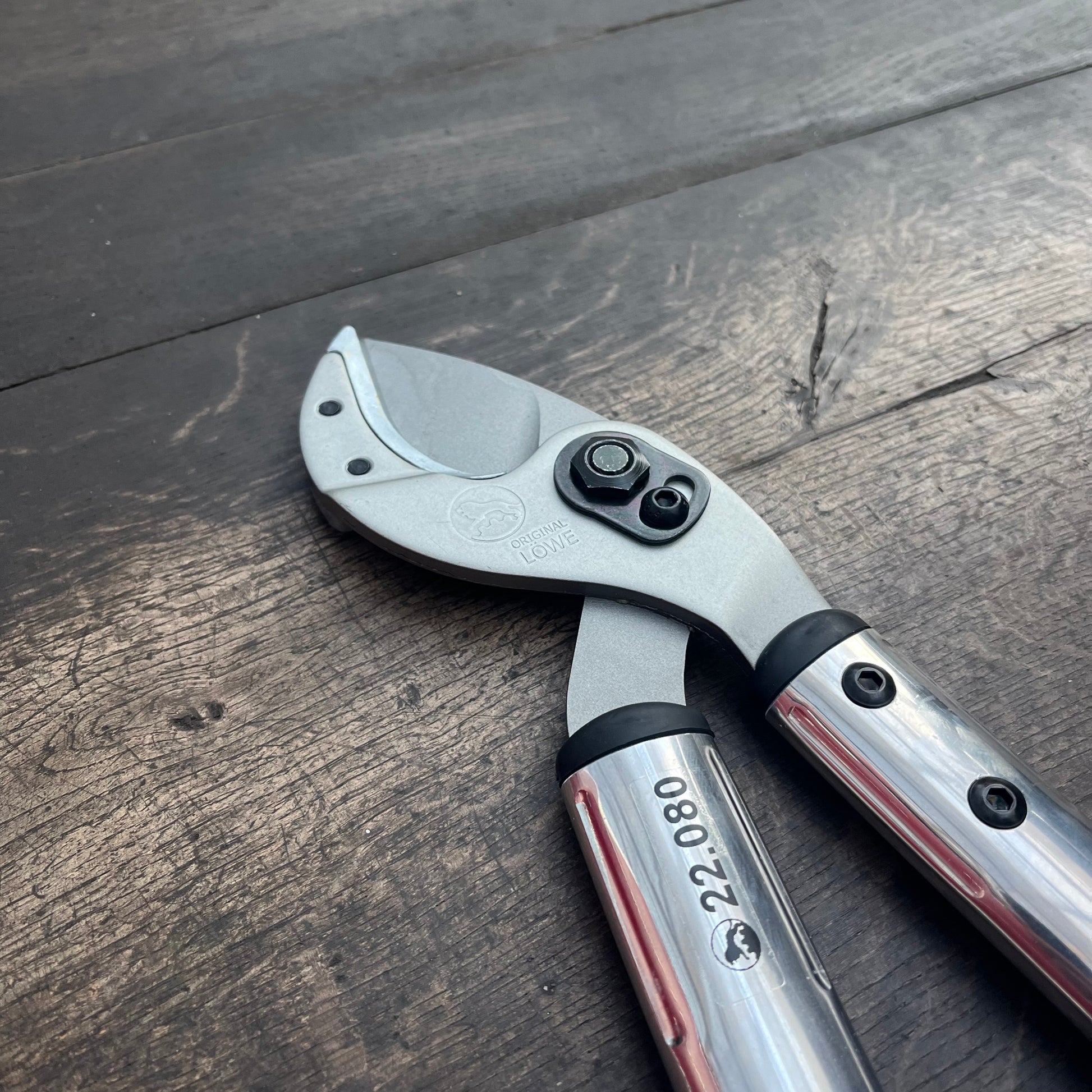 Nahaufnahme der Astschere Löwe Astschere 22 (80 cm) in Silber und Schwarz, mit einer robusten Metallklinge auf dunklem Holz. Ideal für die Gartenpflege. Die nummerierten Griffe und die sichtbaren Schrauben ermöglichen ein präzises Arbeiten beim Baumschnitt. Marke: Löwe.