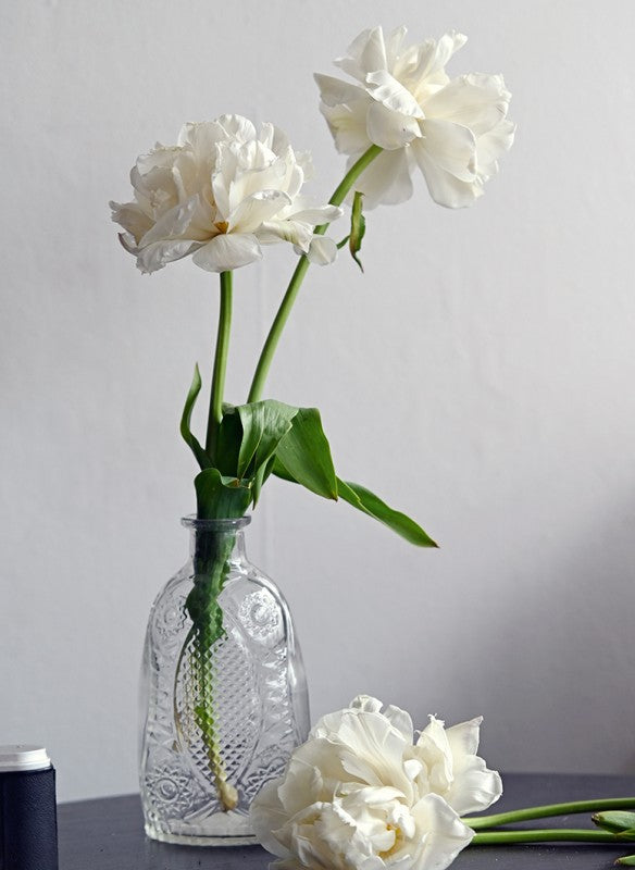 Eine Vase aus klarem Glas mit verschlungenem Muster hält zwei weiße Pfingstrosen mit grünen Stielen und Blättern, die das reine Tulpenglück des Callwey Verlags unterstreichen. Eine weiße Pfingstrose liegt neben der Vase auf einer dunklen Fläche vor einem schlichten hellgrauen Hintergrund.