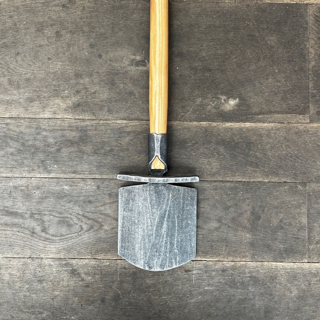 Der Krumpholz Damenspaten 'Elke' (85cm) mit Holzgriff liegt flach auf dunklem Holzboden, das Spatenblatt zeigt nach unten und der Griff ragt aus dem Bild heraus.
