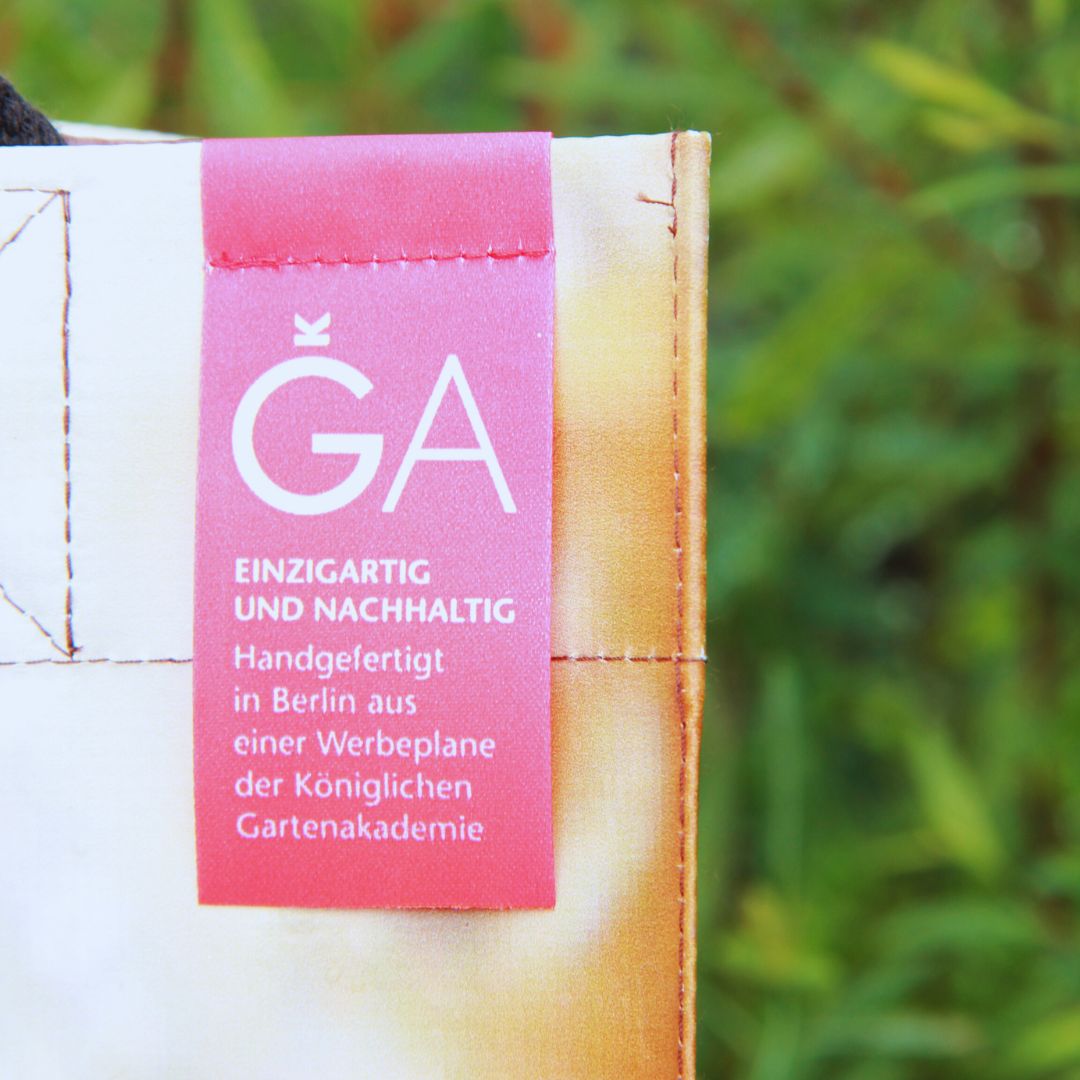 Nahaufnahme eines Stoffanhängers mit deutschem Text auf der Upcycling KGA Tasche "Torf" der Königlichen Gartenakademie, der besagt, dass es sich um ein Unikat handelt, das in Berlin aus einem einmaligen Werbebanner handgefertigt wurde, vor einem unscharfen grünen Hintergrund.