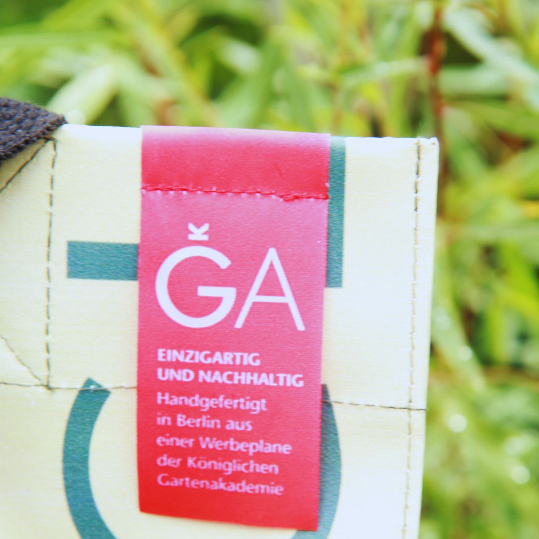 Eine Nahaufnahme eines Unikat-Taschen-Etiketts mit deutschem Text, das auf dem cremefarbenen Upcycling KGA Shopper 'Orange' der Königlichen Gartenakademie angebracht ist. Unscharfes grünes Laub erscheint im Hintergrund.