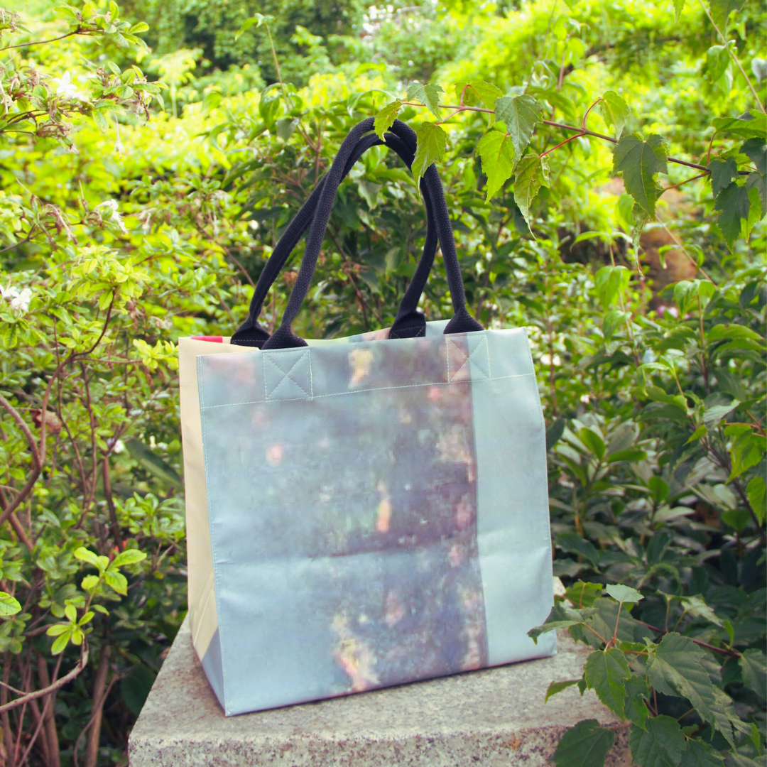 Die Upcycling KGA Tasche 'Sand' der Königlichen Gartenakademie mit schwarzen Griffen ruht auf einer Steinfläche, umgeben von üppigen Grünpflanzen in einem Außenbereich.