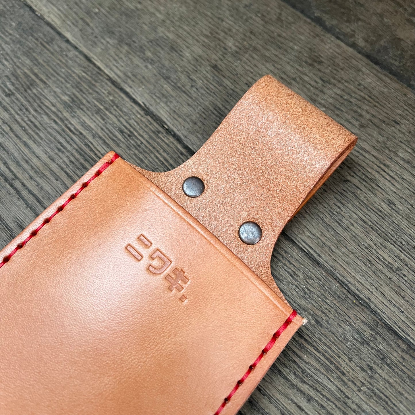 Ein hellbraunes Sägenholster von Niwaki mit roten Nähten und zwei Metallnieten ruht auf Holz. Das Holster für eine Klappsäge hat eine gestempelte Markierung mit japanischen Schriftzeichen.