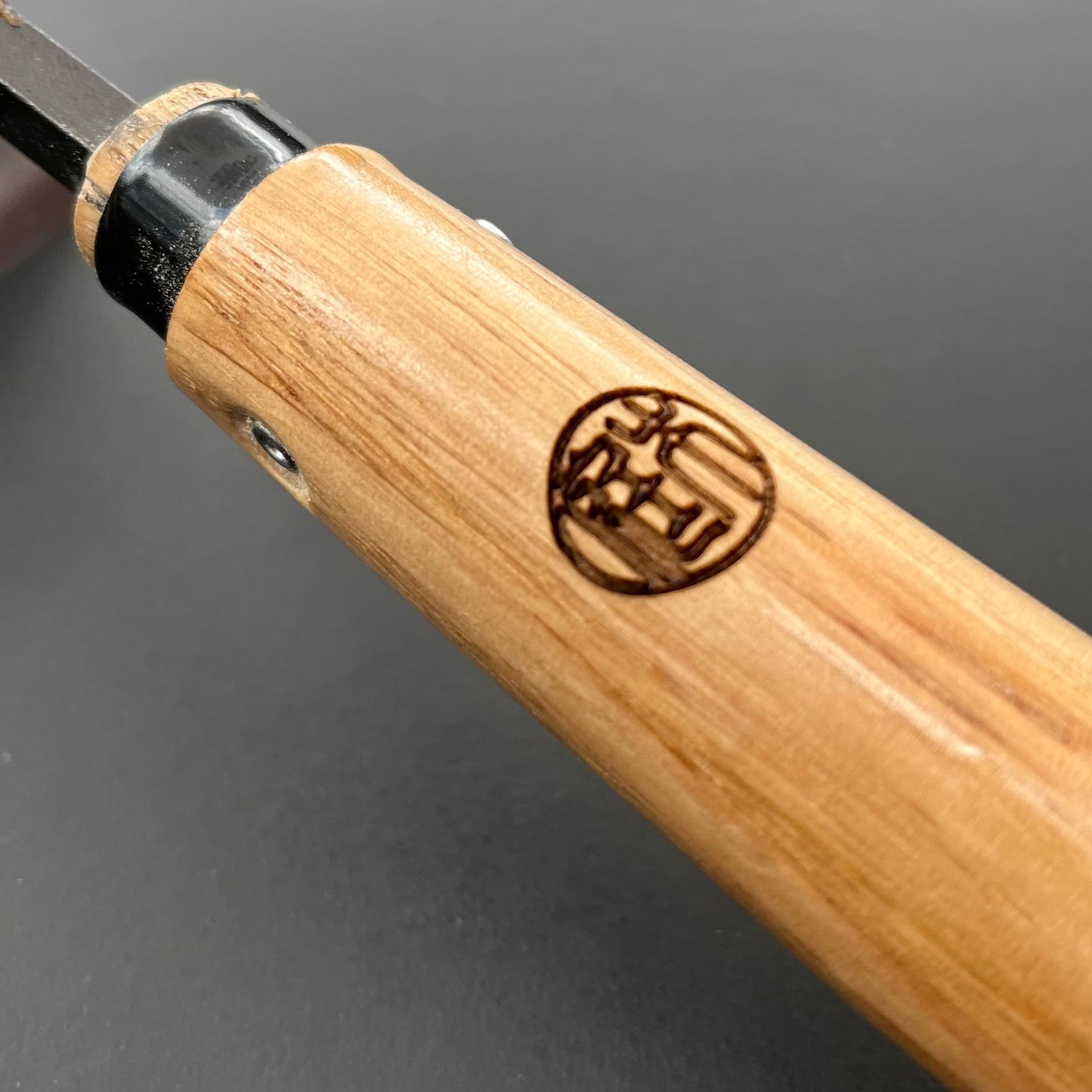 Eine Nahaufnahme der Niwaki Unkrauthacke von Niwaki. Sie verfügt über einen Holzgriff mit einem runden Logo in japanischen Schriftzeichen und ein schwarzes Metallende. Das ergonomische Design ermöglicht müheloses Unkrautjäten vor einem schlichten, dunklen Hintergrund.
