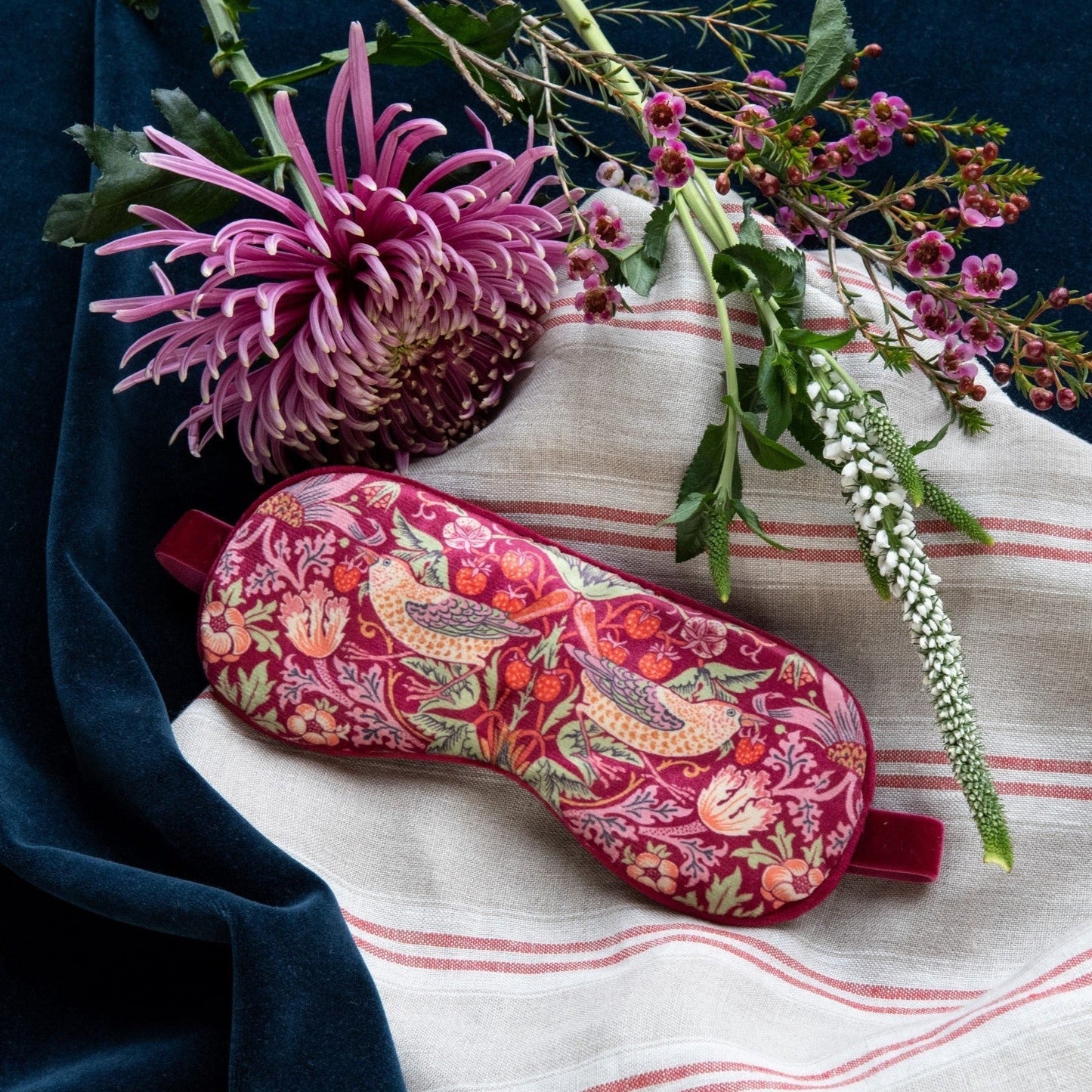 Die William Morris at Home Schlafmaske 'Strawberry Thief', mit getrocknetem Lavendel gefüllt, liegt auf gestreiftem Stoff, umgeben von violetten und weißen Blumen auf dunkelblauem Hintergrund.